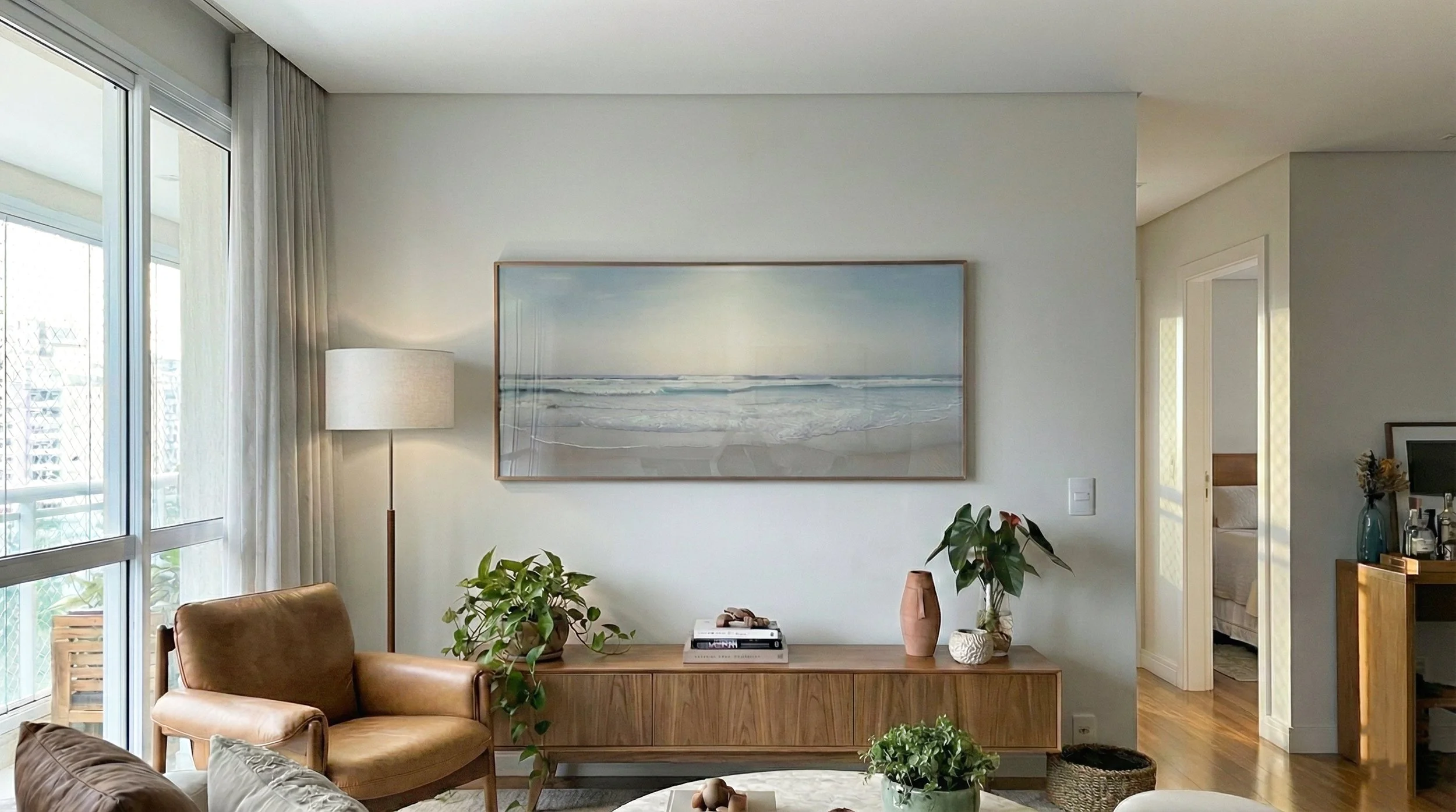 Sala de estar com aparador de madeira clara, luminária acesa e plantas ao lado de um quadro de mar com horizonte suave. Fotografia de Pedro Ferr usada em projetos de interiores que buscam leveza, luz natural e estética equilibrada.