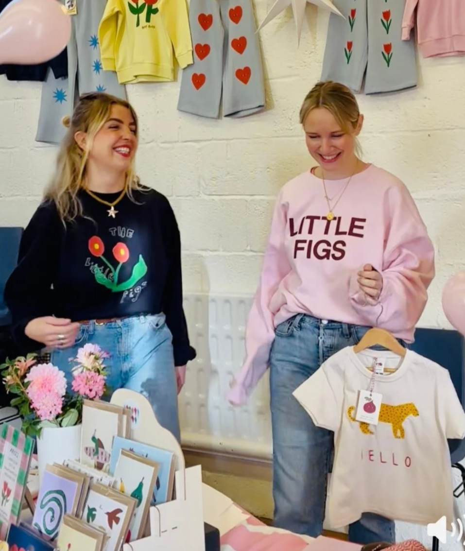 Two women smiling and talking at a craft or clothing booth with decorated Children's T-shirts hanging on the wall behind them, including one pink shirt with 'LITTLE FIGS' and another with a giraffe and 'HELLO'.