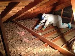 Person wearing protective clothing inspecting attic insulation between wooden beams.