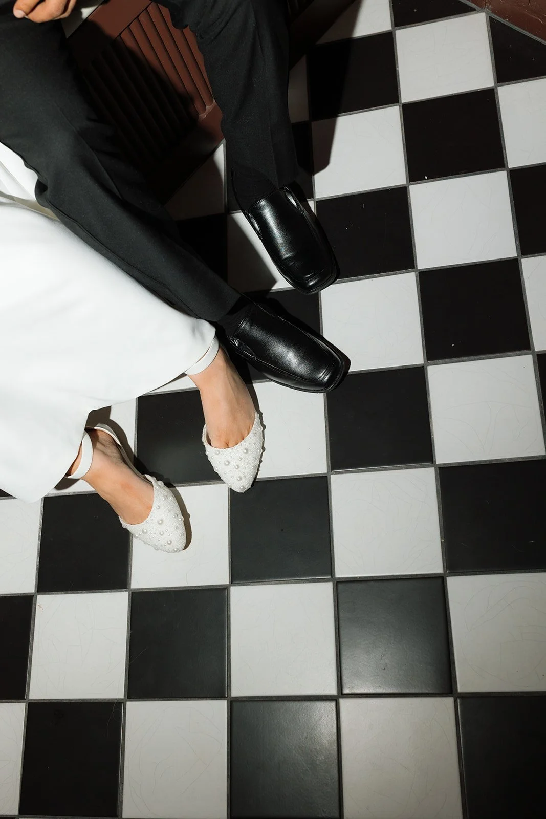 Close-up of two people sitting on a black and white checkered floor, wearing formal shoes and clothing, showing only their legs and feet.