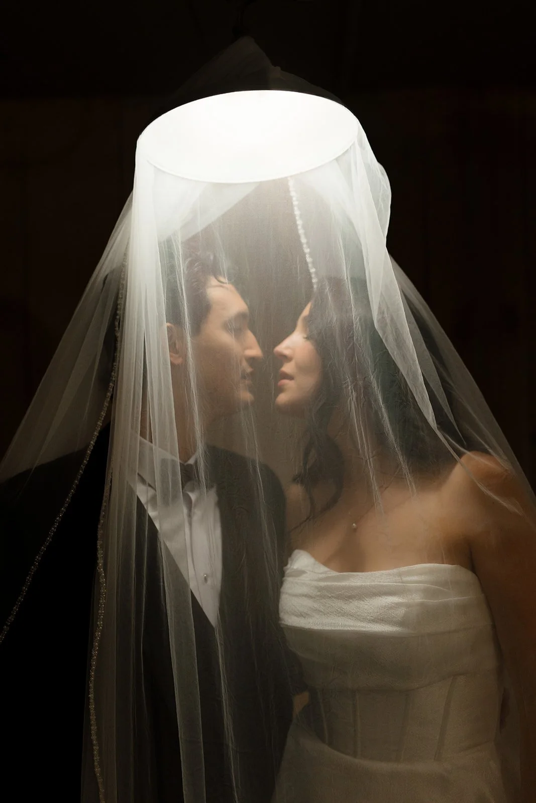 A bride and groom facing each other under a bridal veil with a lamp above them