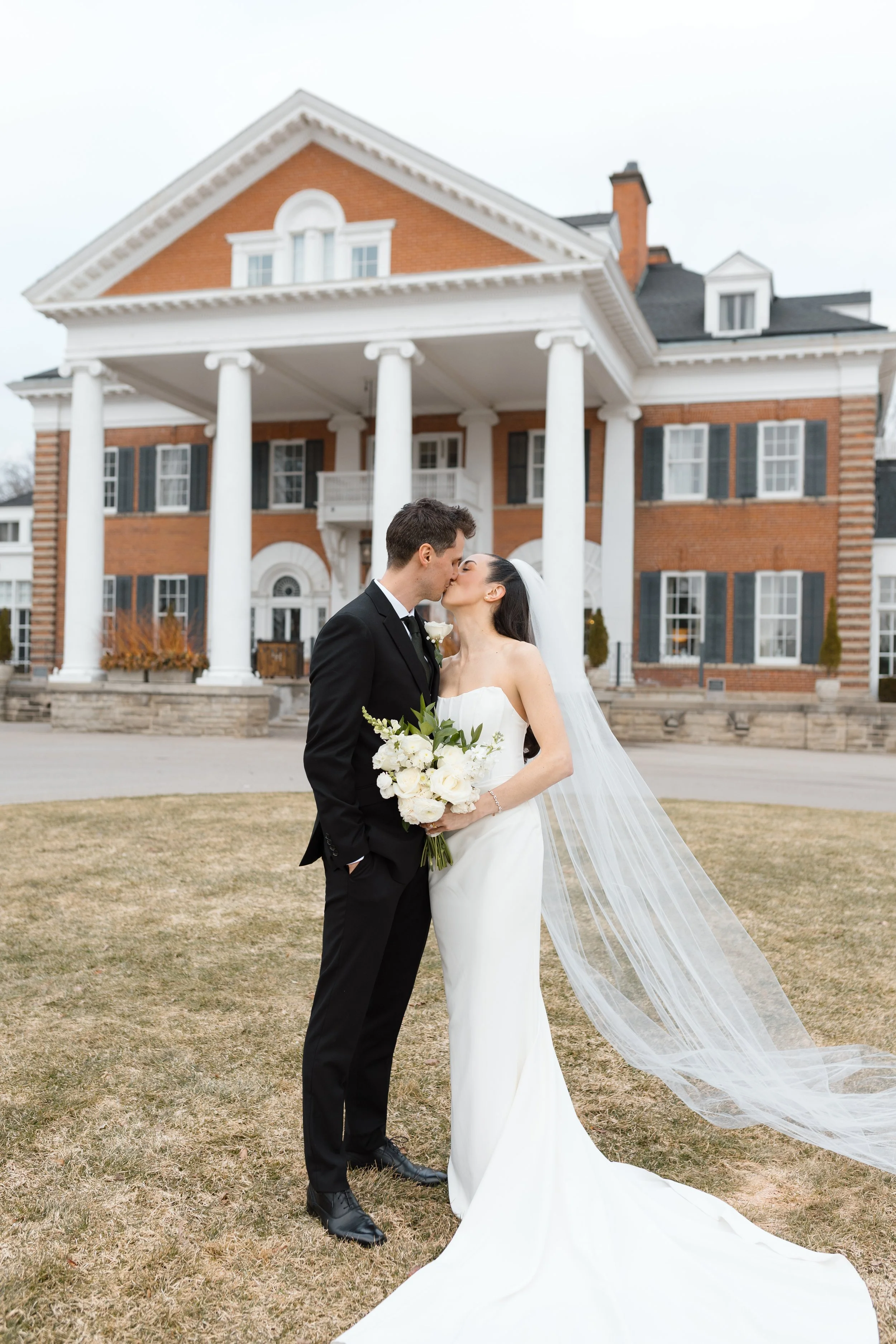 A bride and groom in wedding attire sitting on the grass, smiling and petting a golden retriever dog dressed in a tuxedo, during a wedding outdoor ceremony with trees in the background.