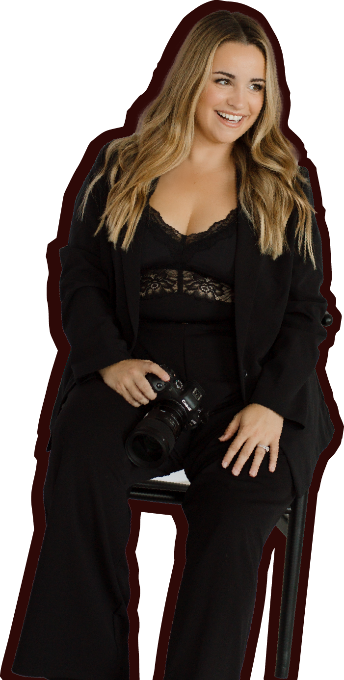 A smiling woman with long wavy blonde hair, wearing a black lace top and black blazer, sitting on a chair and holding a Canon camera in her right hand.