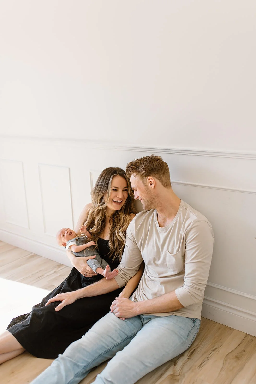 A smiling woman holding a baby, sitting next to a man, all sitting on the floor against a white wall.