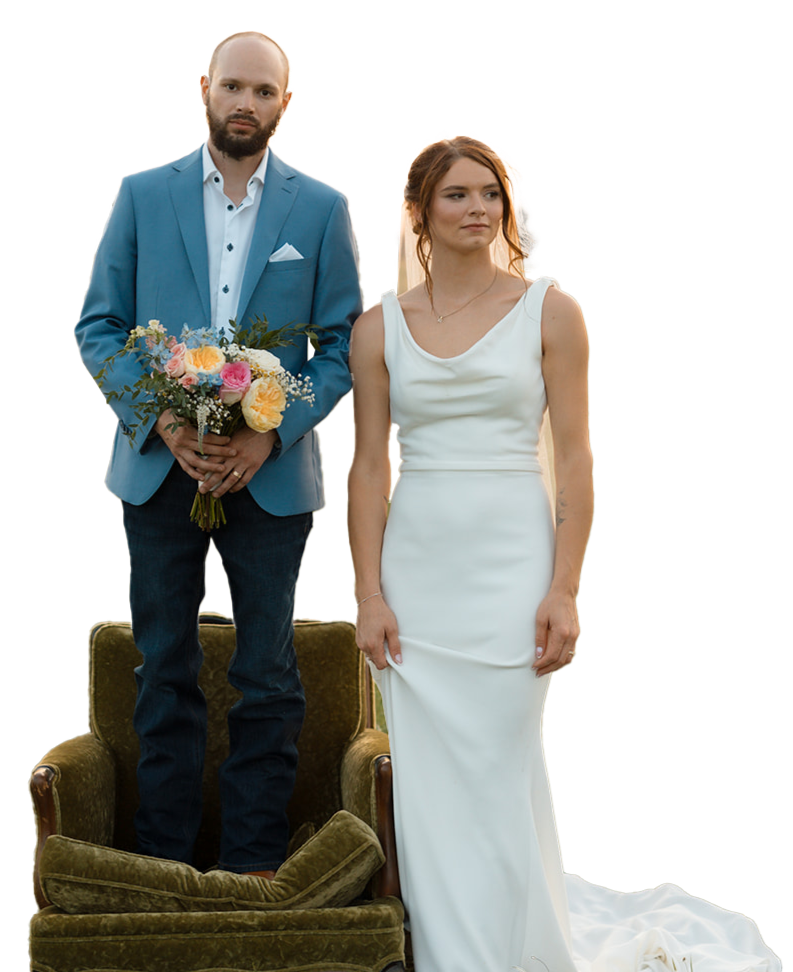 A man in a blue suit holding a bouquet of flowers standing on a vintage green armchair, next to a woman in a white dress standing on the floor.