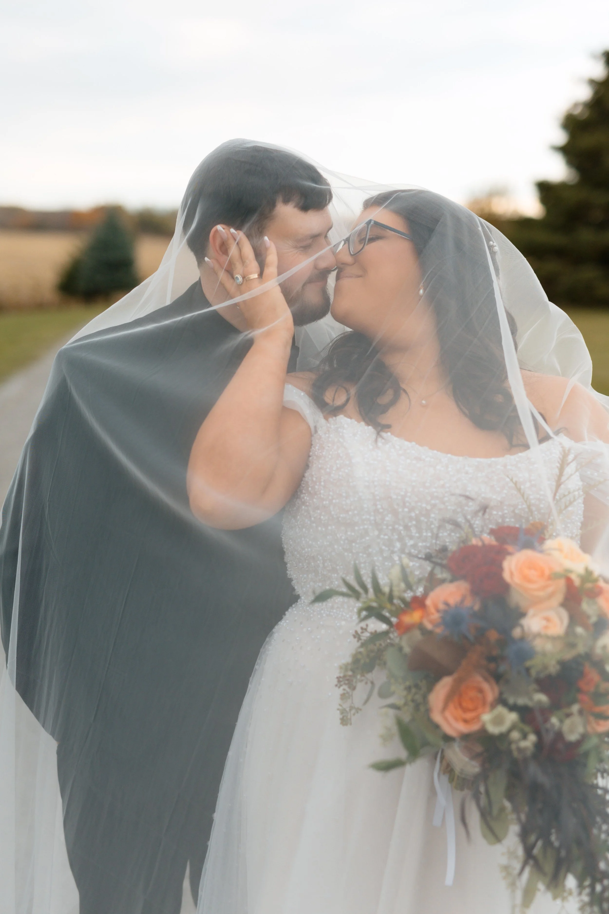 A bride and groom under a wedding veil sharing a moment outdoors, with trees and a cloudy sky in the background.