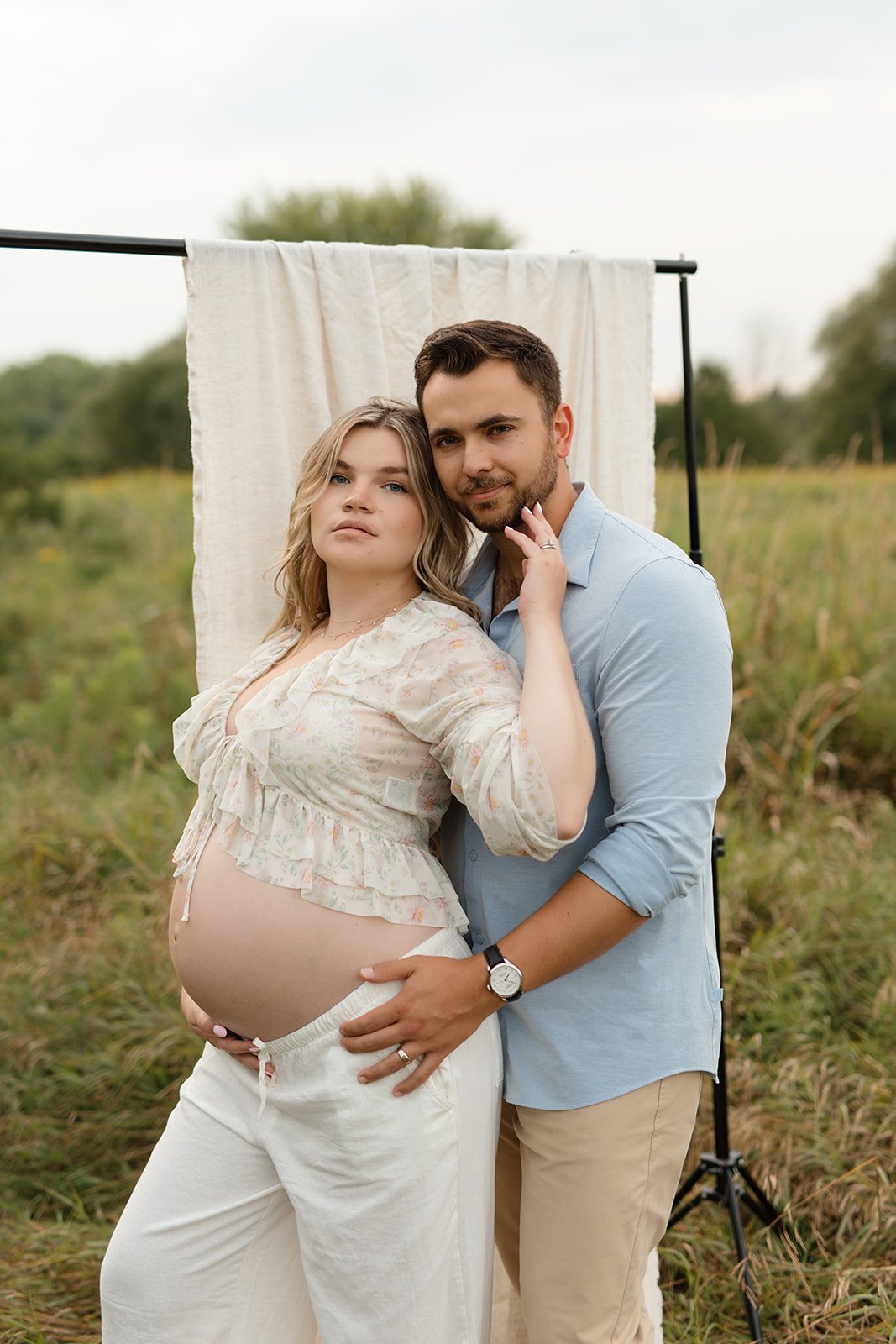 Pregnant woman and man standing together outdoors with a white fabric backdrop, embracing each other, in a grassy field.