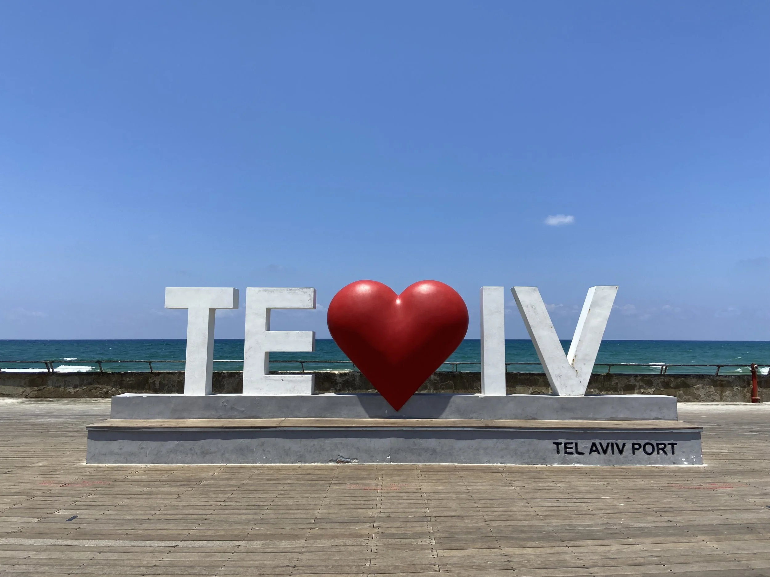 Sign spelling 'I ❤️ TV' with a red heart in place of 'love', situated at Tel Aviv Port, overlooking the sea under a clear blue sky.
