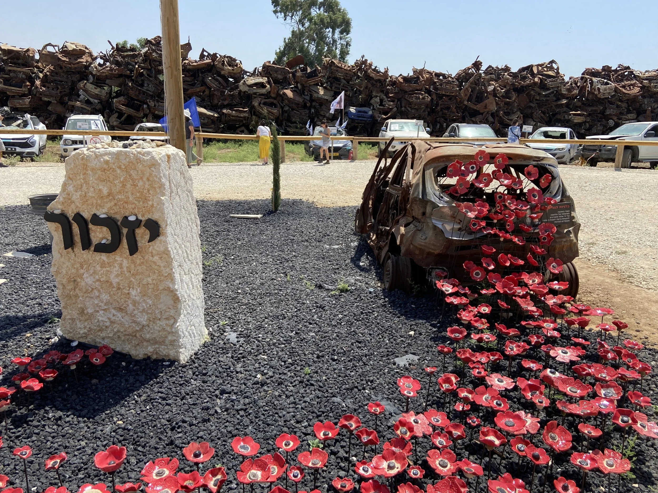 Memorial site with a stone marker inscribed with Hebrew script, an array of red artificial poppies on black stems, and a wrecked, burnt-out car with more poppies attached. In the background, a wall of rusted cars and several people observing the site.