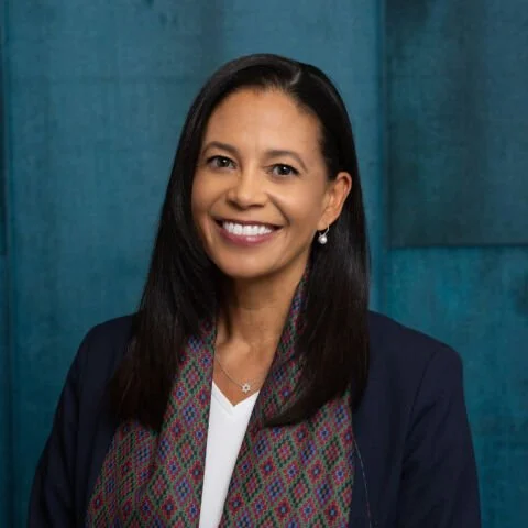 Headshot of a woman with long dark hair, wearing a dark blazer, patterned scarf, and a white top, smiling in front of a blue background.