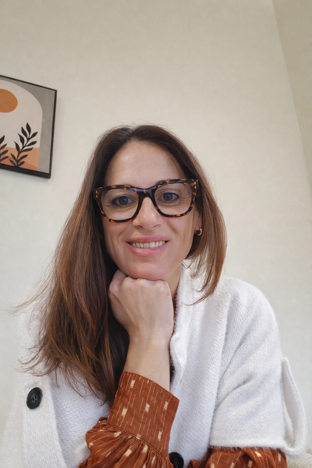 Portrait d'une femme portant des lunettes à monture écaille, avec des cheveux bruns longs, vêtue d'un pull blanc et d'une chemise à manches longues de couleur marron à motifs. Elle pose la tête appuyée sur sa main, souriante devant un mur clair avec une peinture abstraite