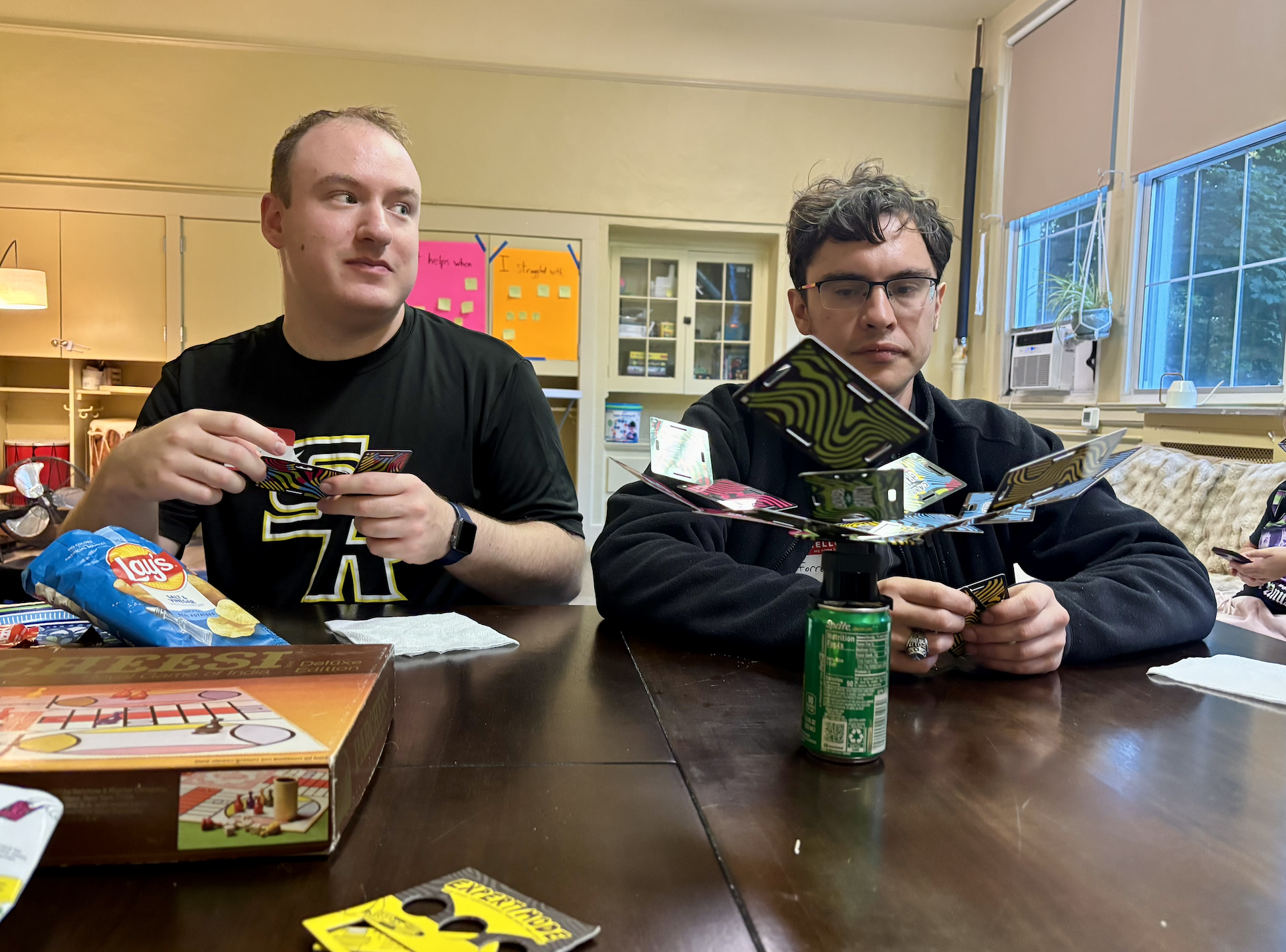 A group of people playing cards around a wooden table with snacks and drinks.