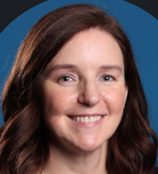Close-up portrait of a woman with long, wavy brown hair smiling in front of a blue background.