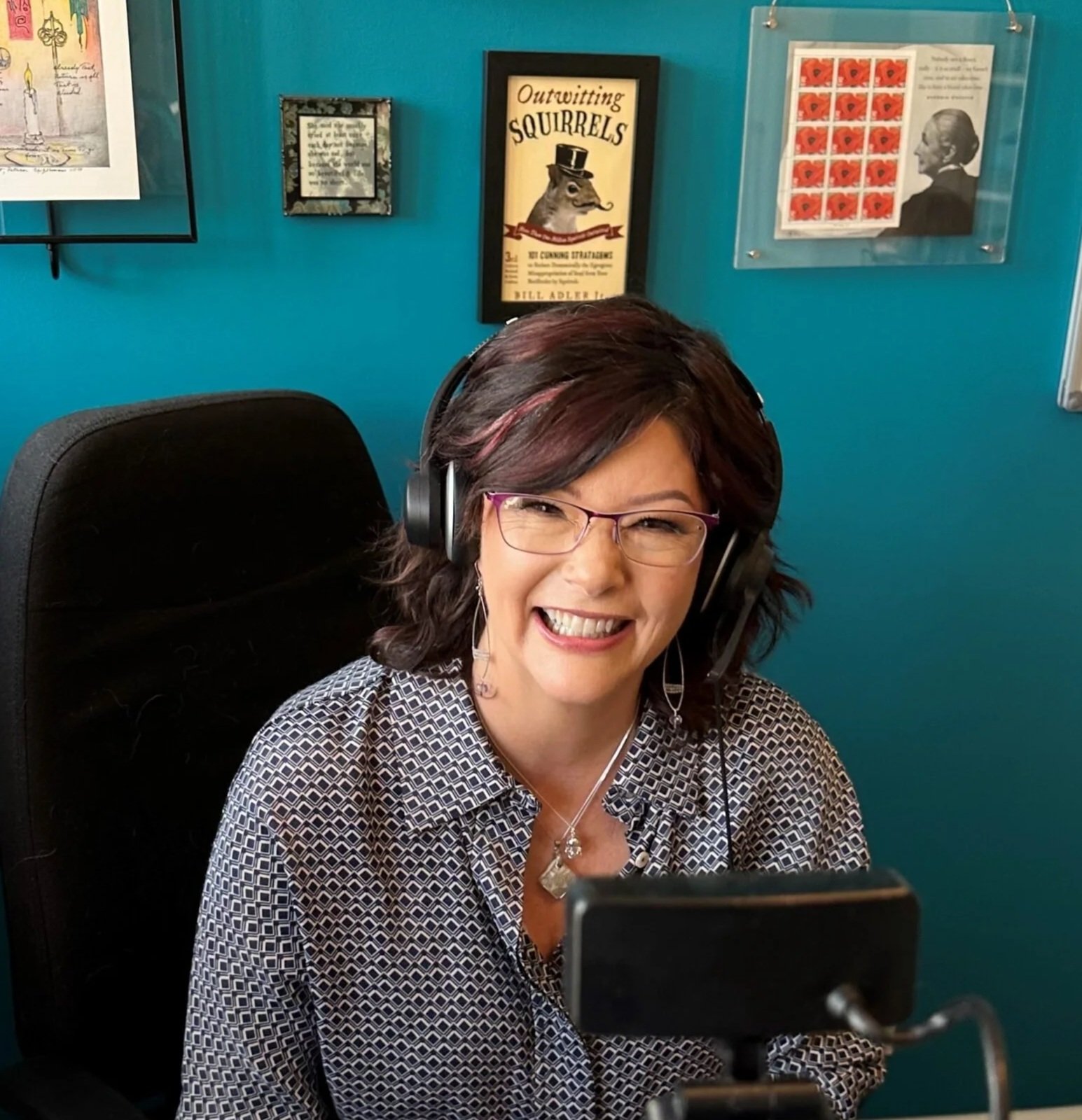 A woman with dark, shoulder-length hair and glasses, smiling, wearing headphones and a patterned blouse, sitting in an office. Behind her are framed posters and artwork on a teal wall.