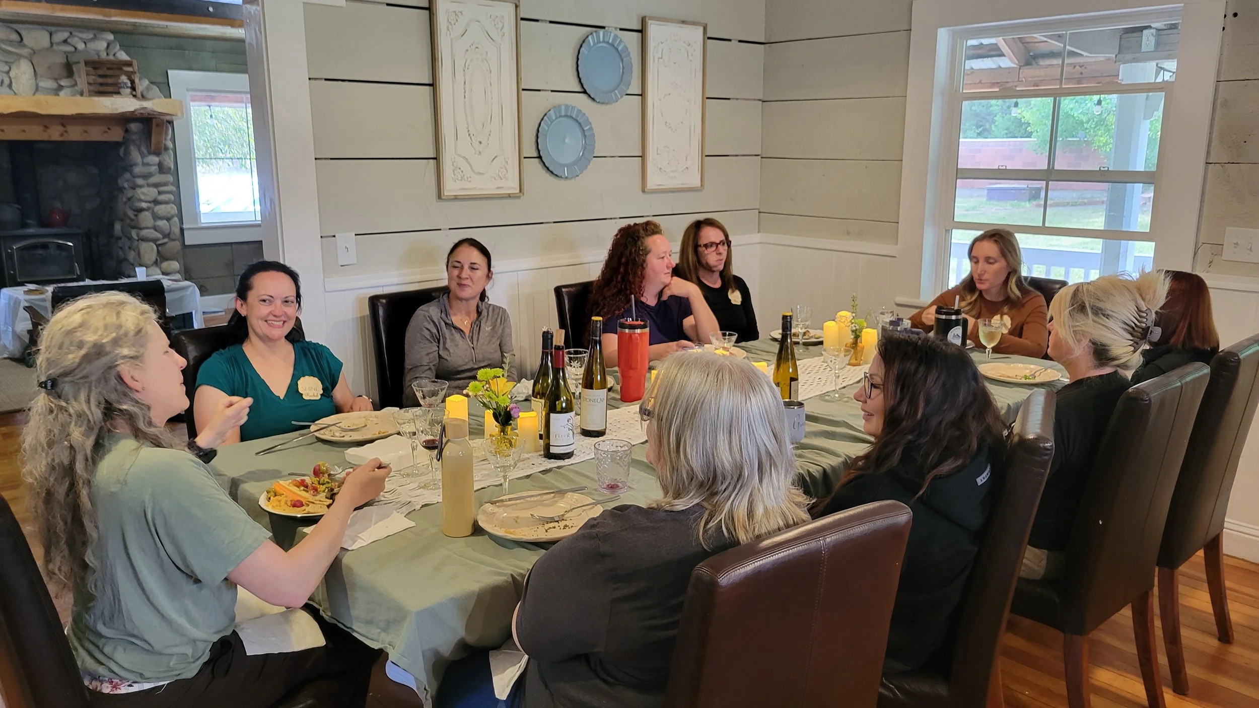 Group of women sitting around a dining table having a meal and conversation inside a house, with a window showing a backyard scene.