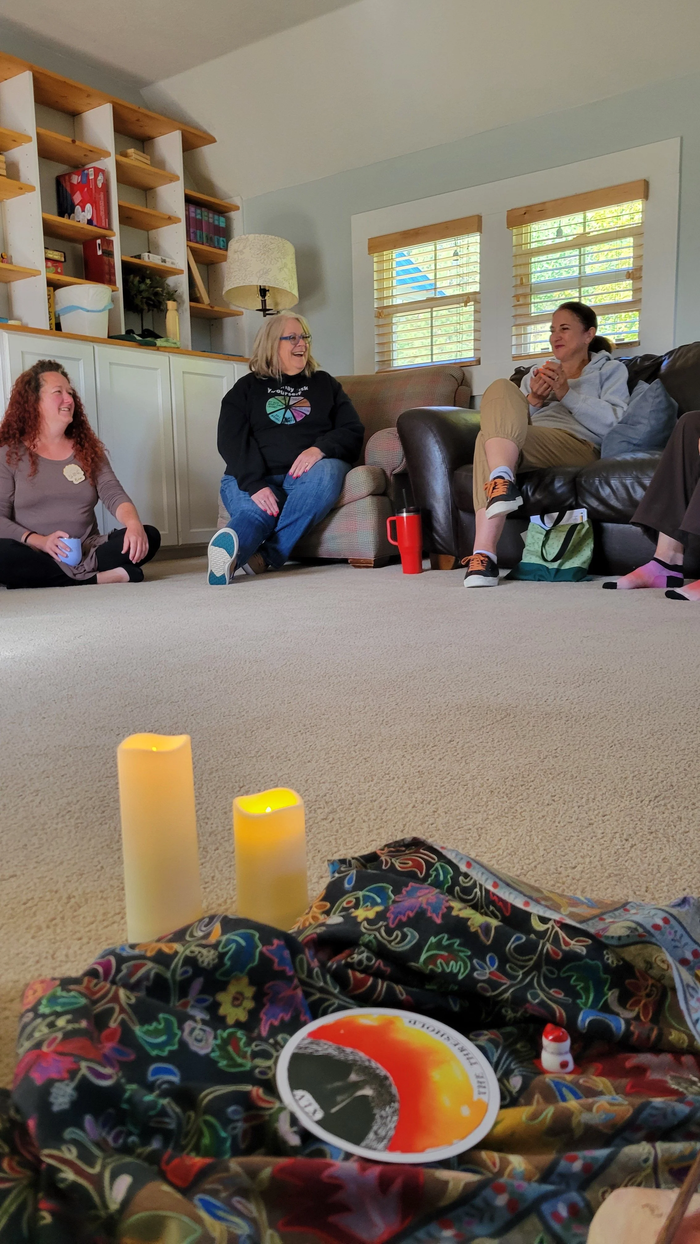 Four women sitting on the floor and on a couch in a living room, smiling and talking. There are candles and colorful cloth on the carpet, with a bookshelf and window in the background.