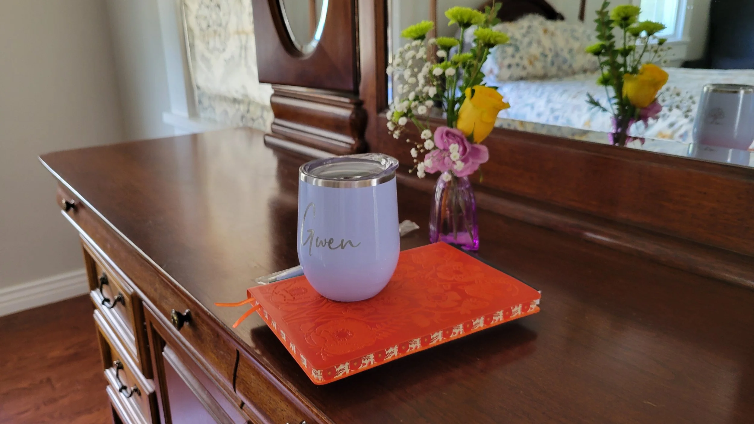 A wooden dresser with a mirror, decorated with a glass vase of flowers, a personalized tumbler with the name Gwen, and a book with an orange cover, in a room with a bed and window.