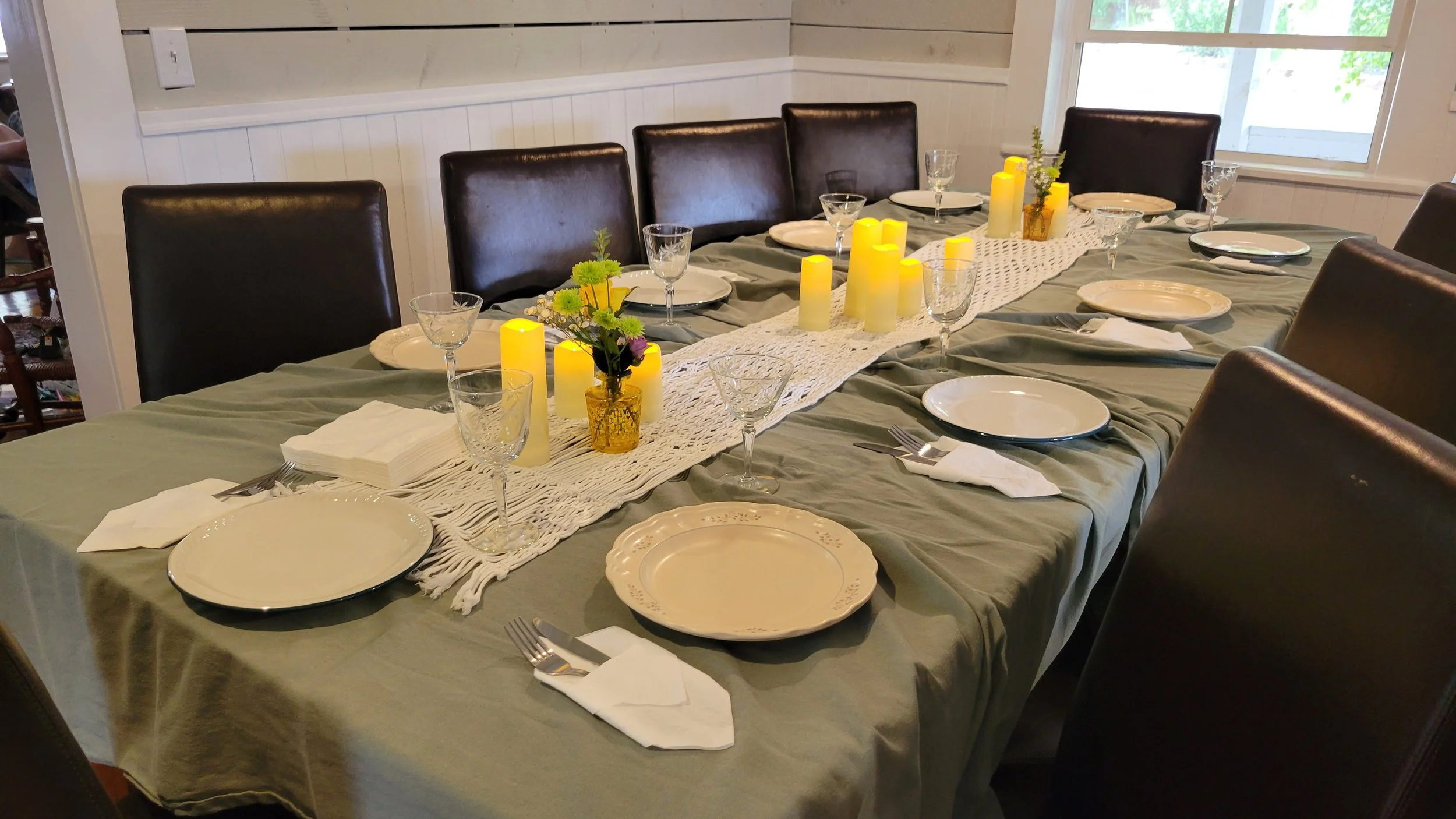 Dining table set for a meal with plates, glasses, napkins, and cutlery, decorated with yellow candles and a small flower arrangement, in a bright room with large windows.