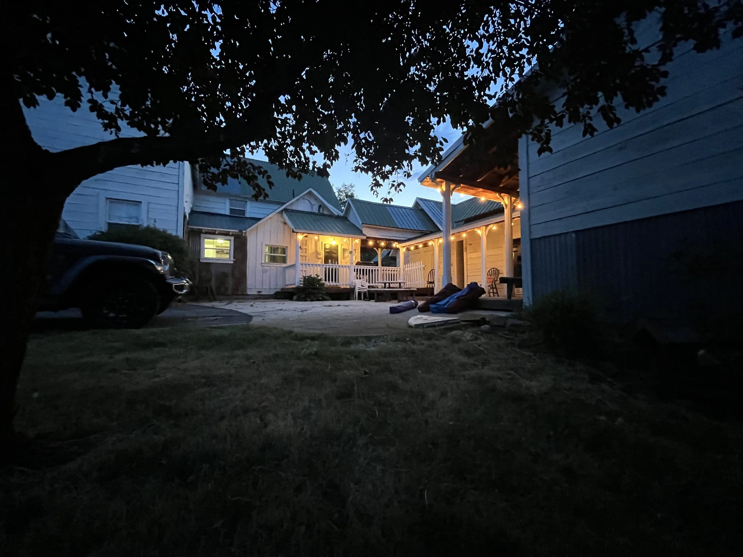 A backyard scene at twilight with a large tree in the foreground, a house with string lights on the porch, and a vehicle parked in the driveway.
