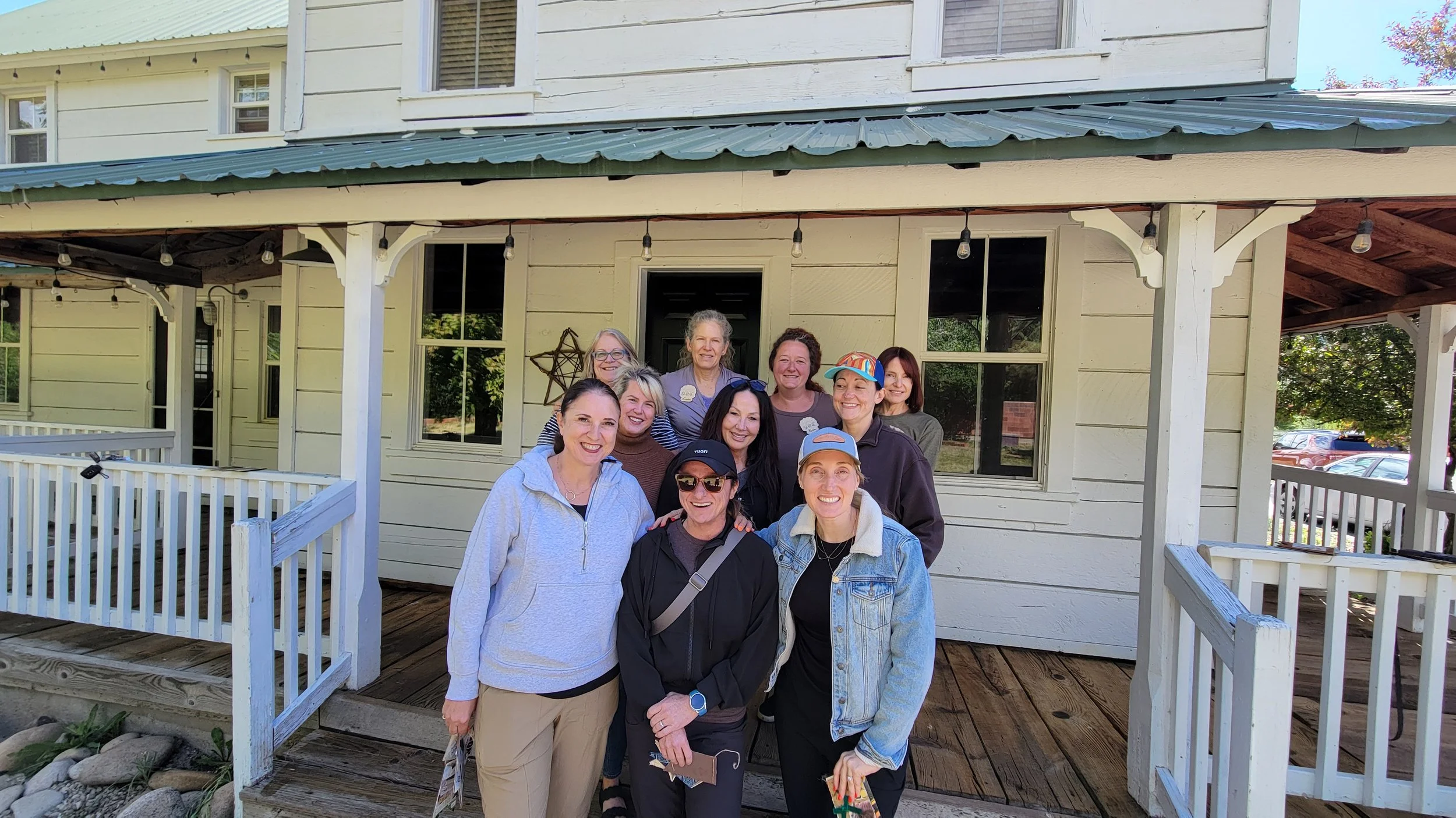 A group of women standing on a porch in front of a yellow house with white trim and string lights hanging from the roof. They are smiling and posing for the photo.