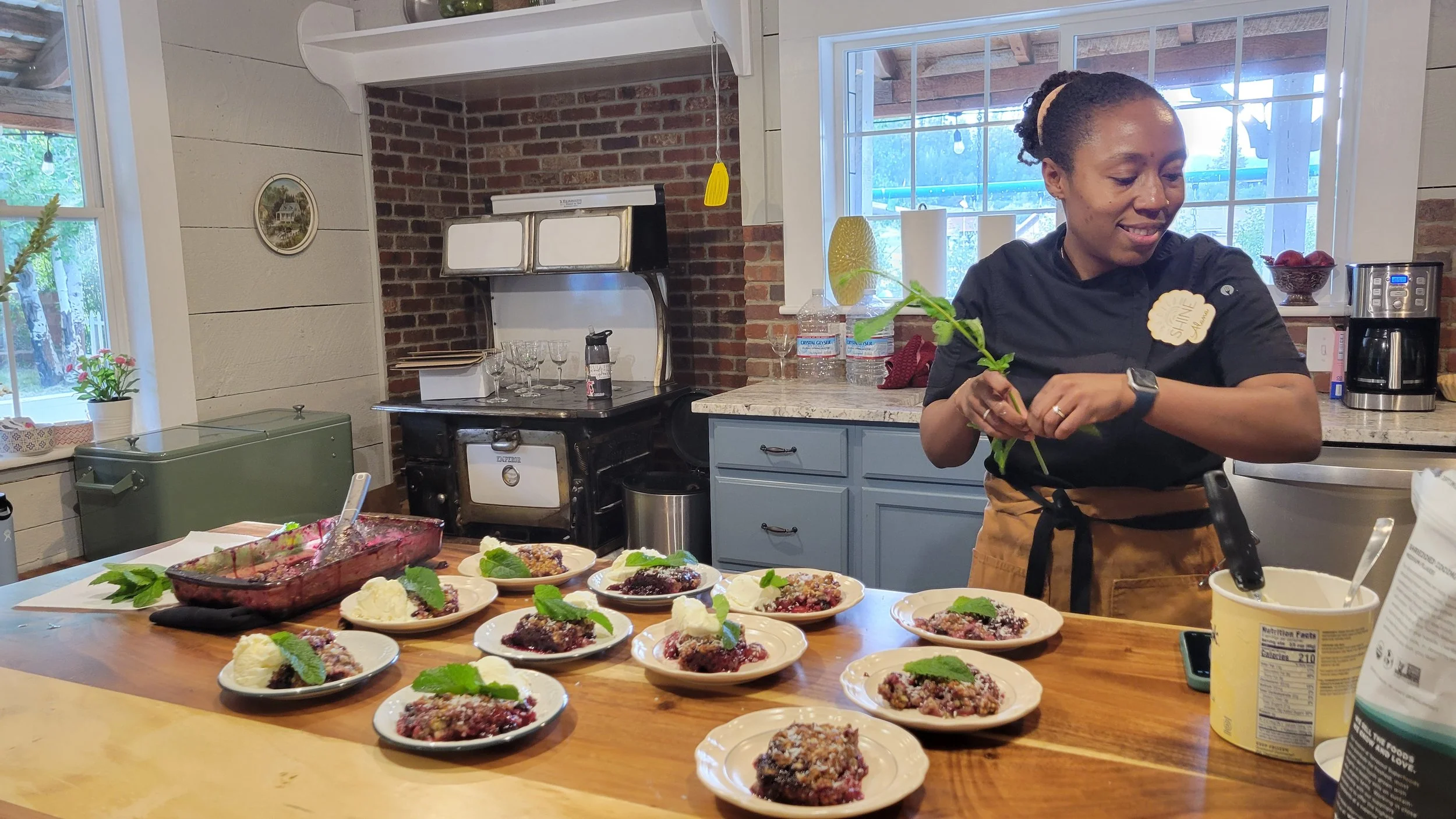A woman preparing dessert at a kitchen counter with plates of berry crumble topped with mint leaves and whipped cream. The kitchen has a rustic style with exposed brick walls, a window, and various appliances.