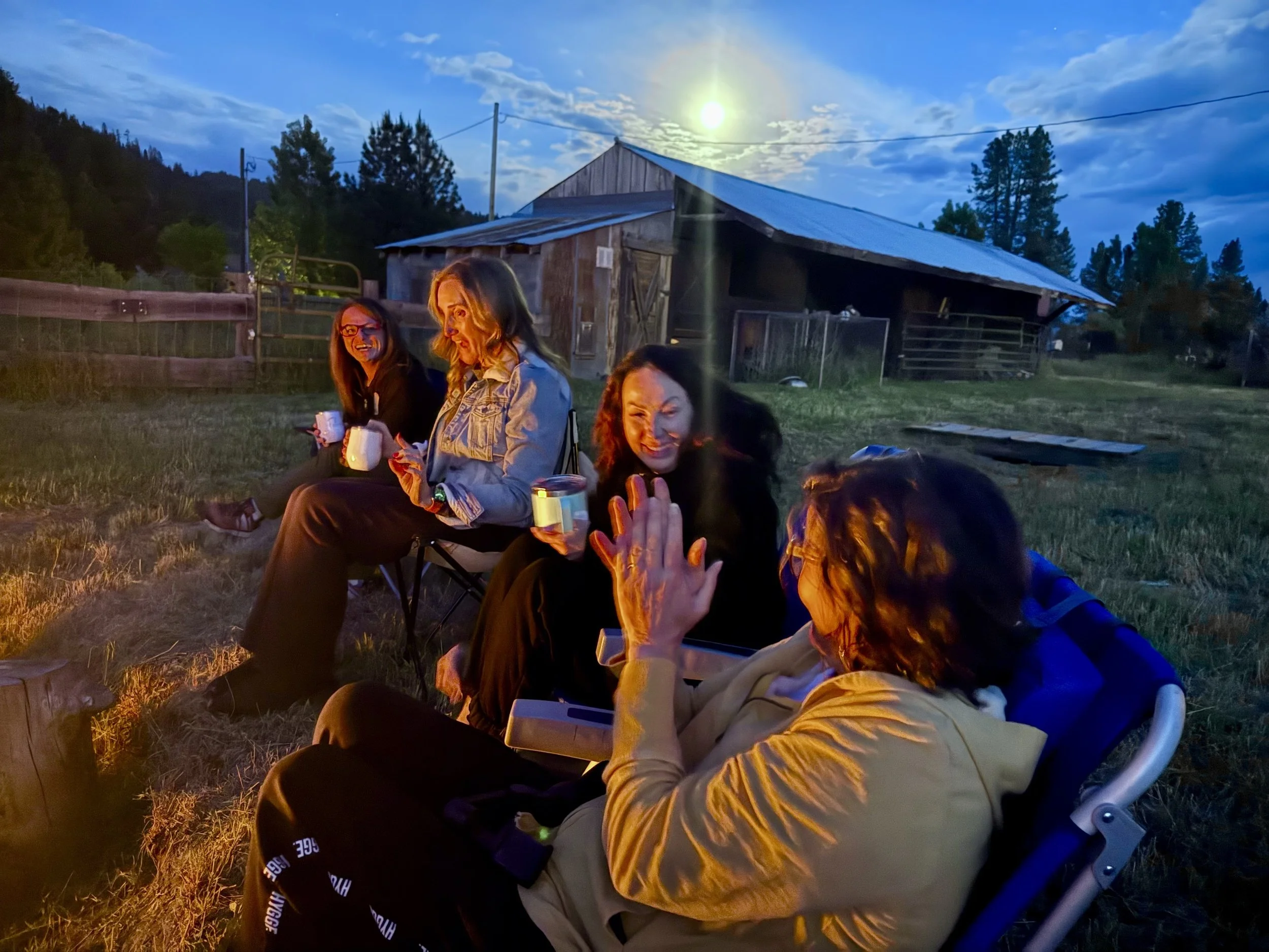 Three women sitting outdoors near a farm building under a bright moonlit sky, enjoying a casual evening together.