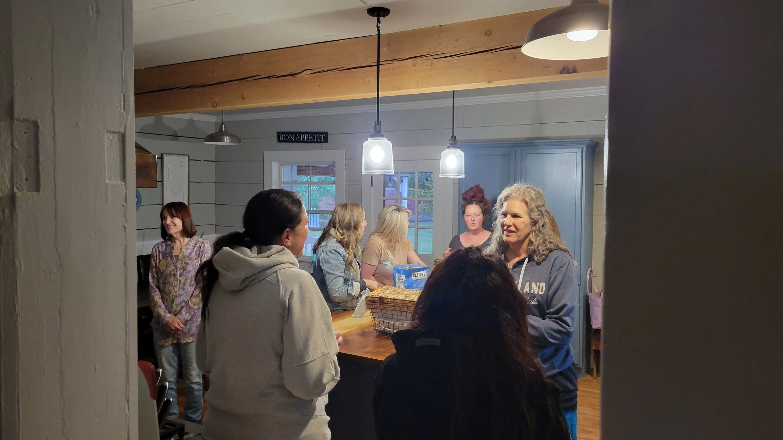 A group of women gathered in a kitchen, engaging in conversation and smiling, with a woman wearing a gray hoodie speaking to others, and a sign that says 'Bon Appetit' hanging on the wall.