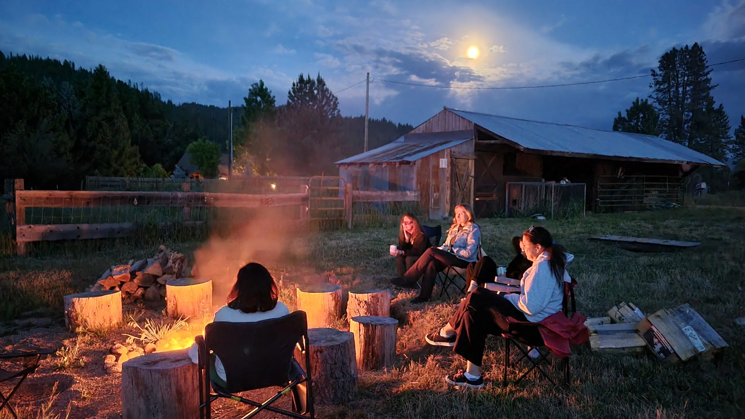 People sitting around a campfire in a rural backyard at dusk, with a barn and trees in the background, and the moon visible in the sky.