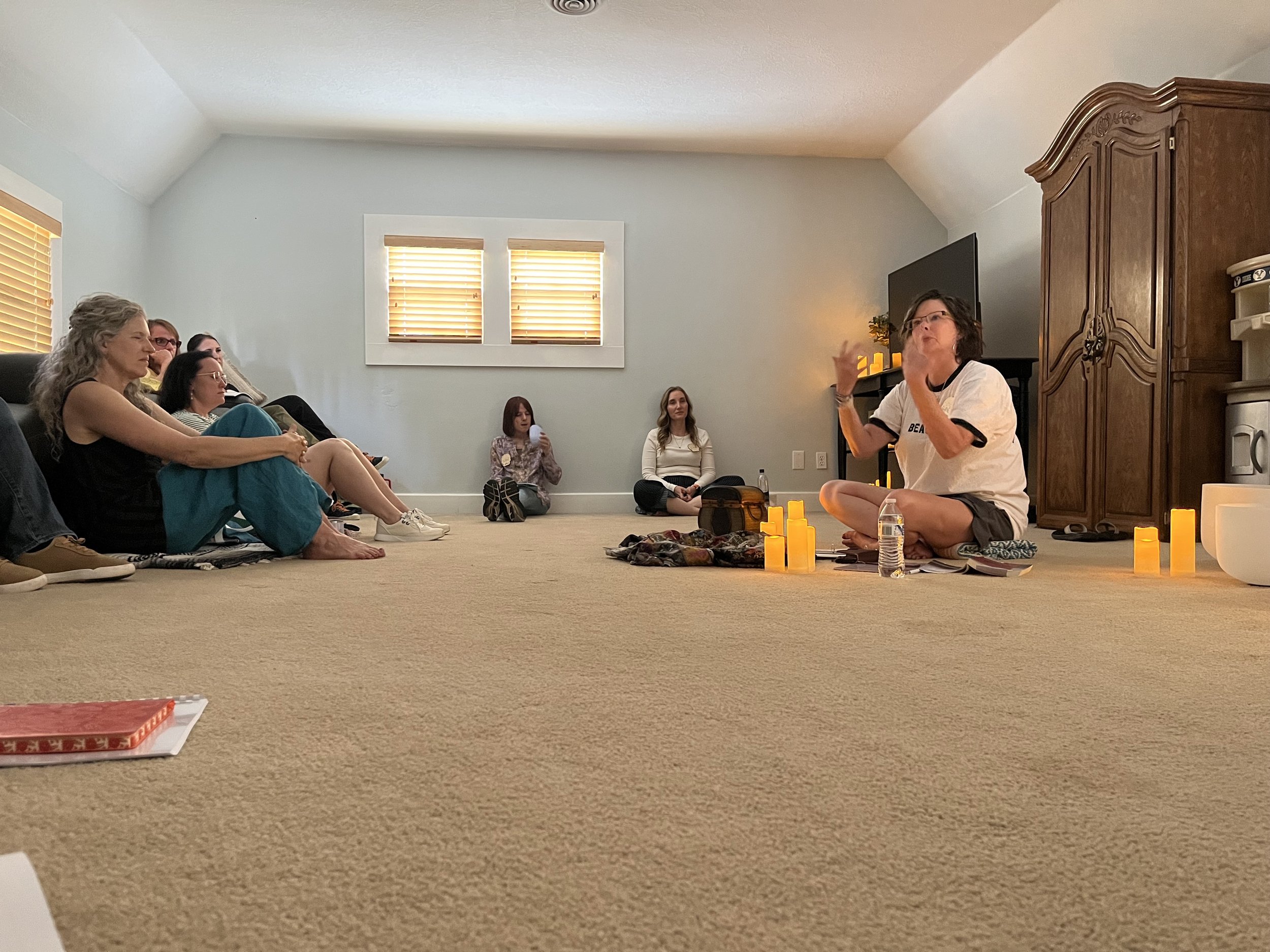 A woman is speaking to a group of people seated on the floor in a living room, with candles arranged in front of her. Others are sitting attentively, and the room has light-colored walls, wooden blinds on windows, and a large wooden cabinet.
