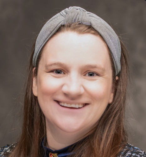 A woman with long brown hair wearing a gray headband and a dark patterned top, smiling at the camera.