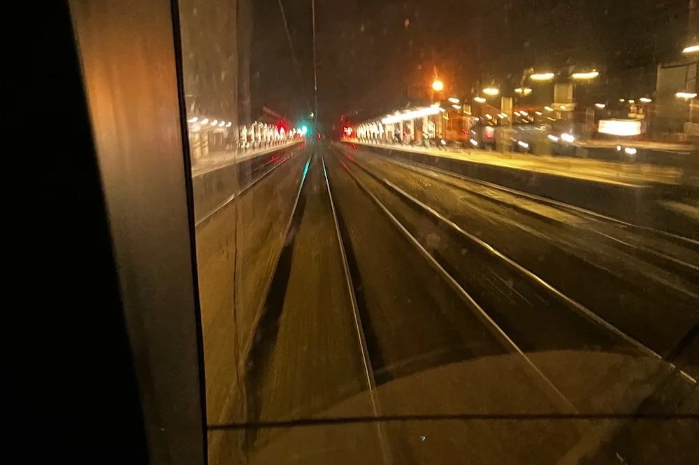View from the driver's cab window of a modern Royal Mail train at night as it speeds through brightly lit Doncaster station's at night. A link to a piece of writing by Dan Richards in memory of his friend, playwrite Oliver Emanuel.
