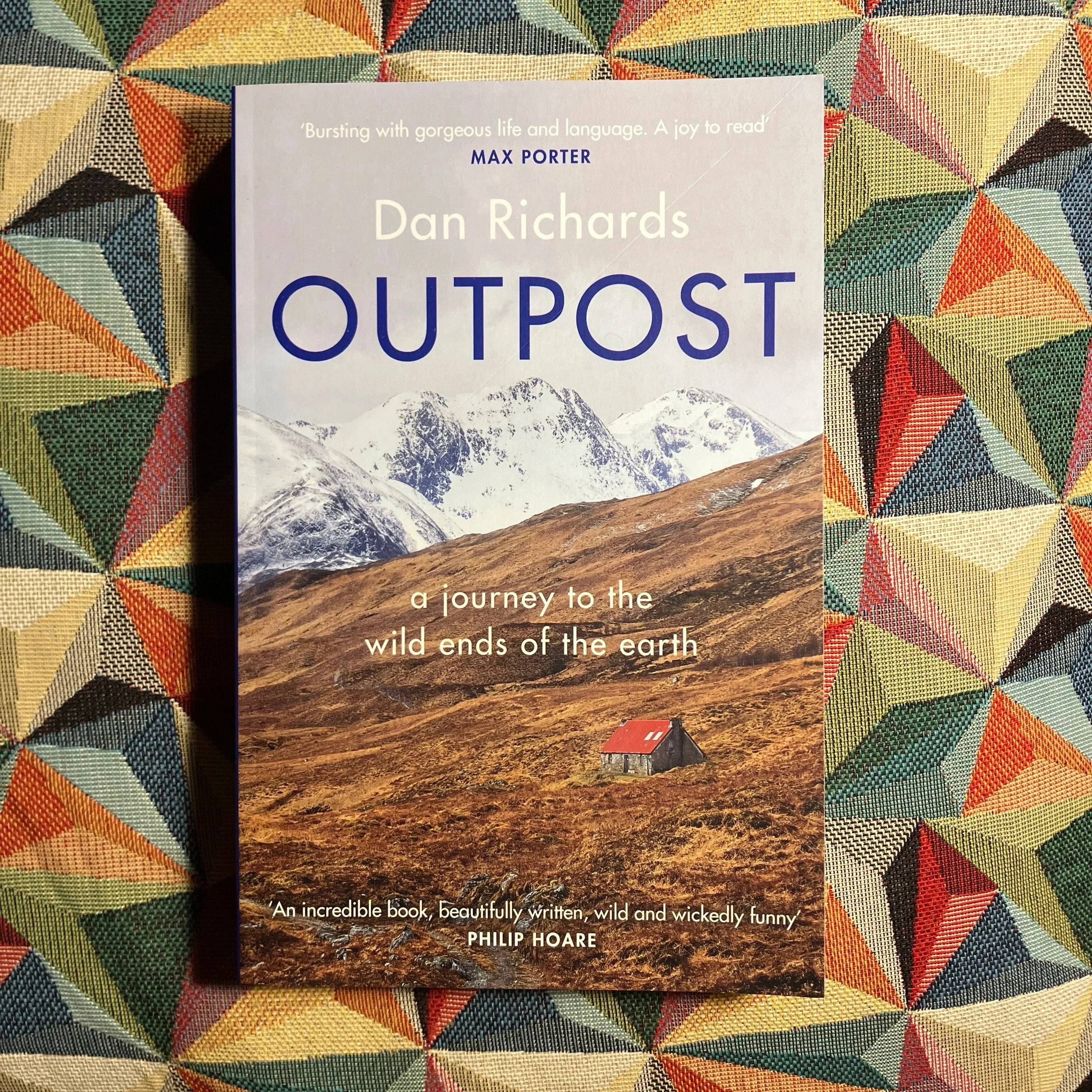 Book titled "Outpost" by Dan Richards on a colorful geometric patterned table, featuring a mountain landscape with snow-capped peaks, a mountainous foreground, and a small house or 'bothy' with a red roof.
Published by Canongate Books in 2019 / 2020