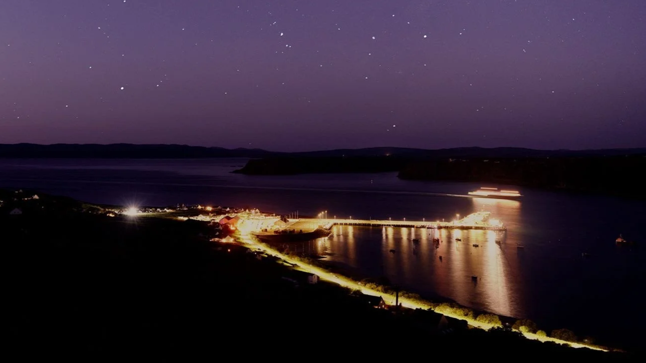 An image from the paperback cover of Overnight a book by writer Dan Richards. A nighttime view of a harbour on the Isle of Skye, Scotland, with lit pathways, calm water reflecting lights, boats anchored, and a starry sky above.