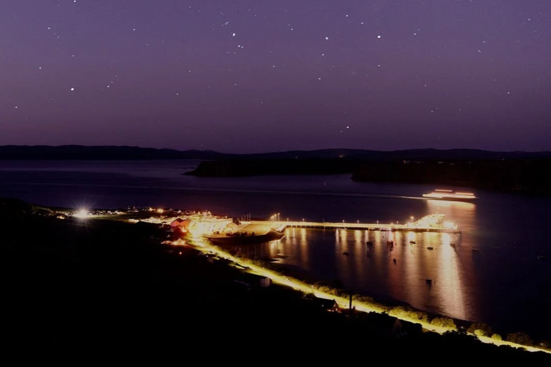Night view of Uig, a coastal town on Skye, Scotland, showing illuminated streets, a pier extending into the sea, calm harbor, a lit-up ferry, and starry sky. The photograph is part of the cover for writer Dan Richards' 2026 paperback, Overnight.