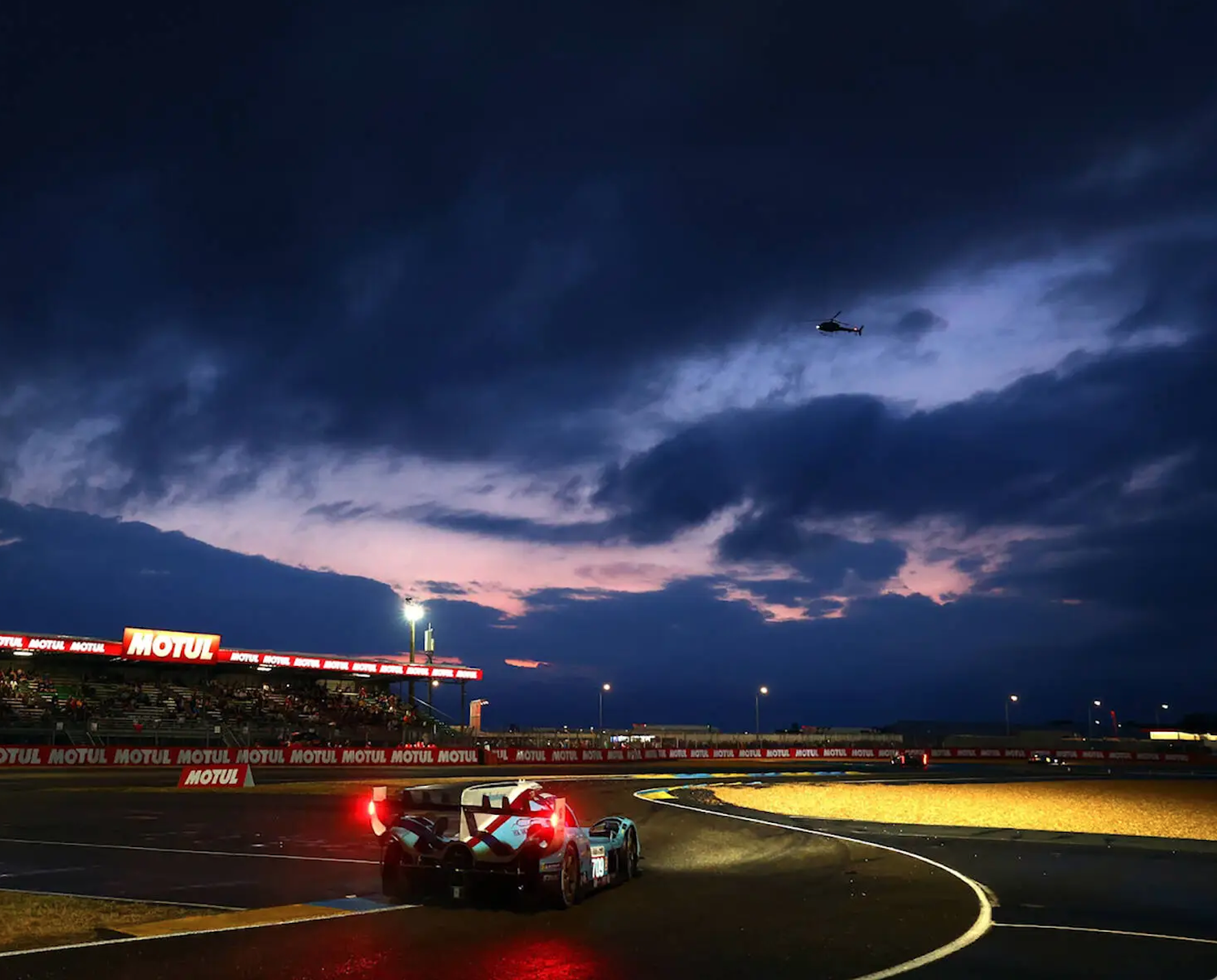 Race car on a track at dusk with a helicopter flying in a cloudy sky and bright lights illuminating the scene. The lead image from a review of Overnight, a 2025 book by Dan Richards, in the Winnipeg Free Press.