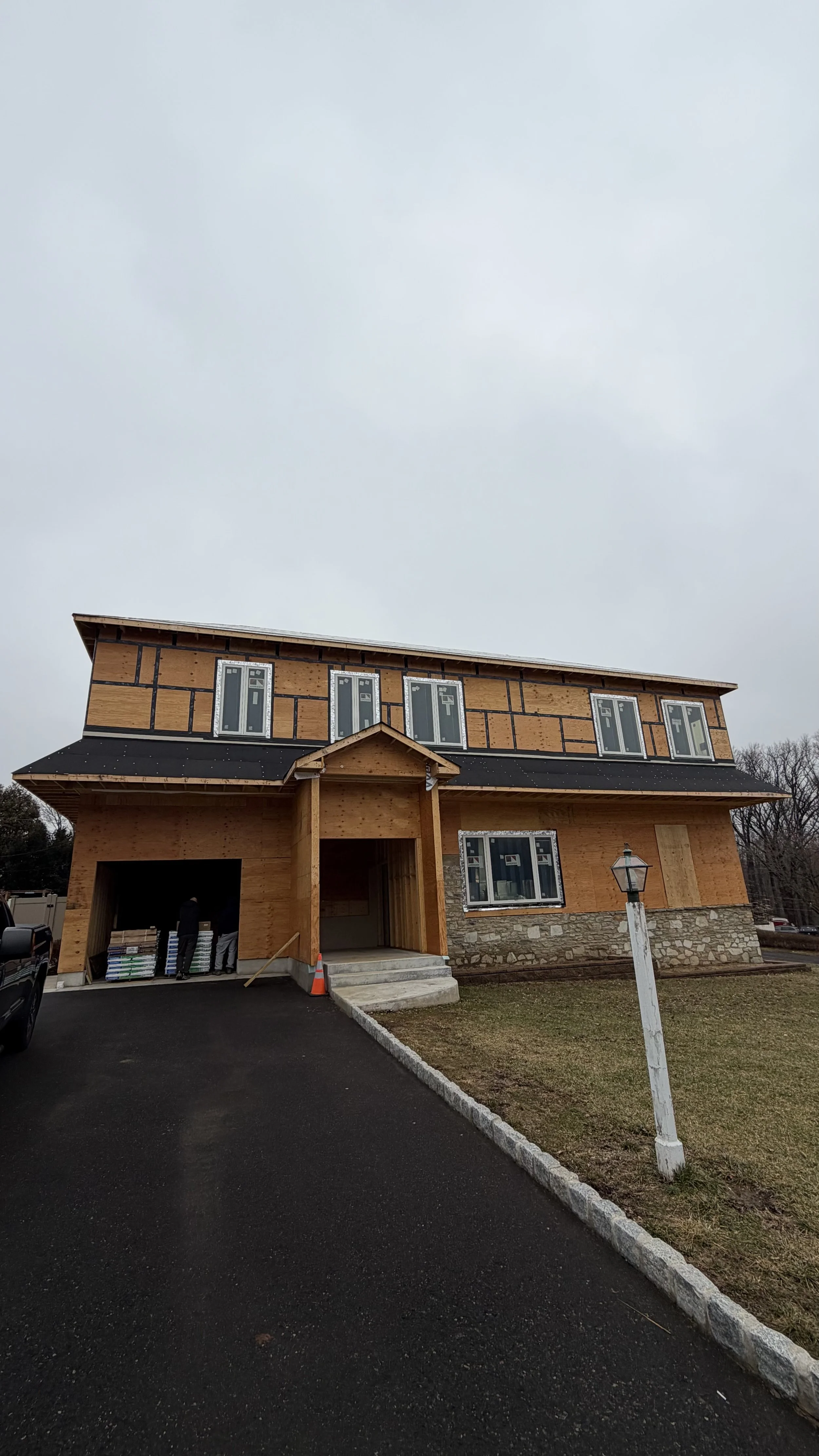 A two-story house under construction with a wood framework and black roofing. The porch is not yet finished, and there are construction materials and workers visible. The sky is overcast, and a lamppost is in the front yard.