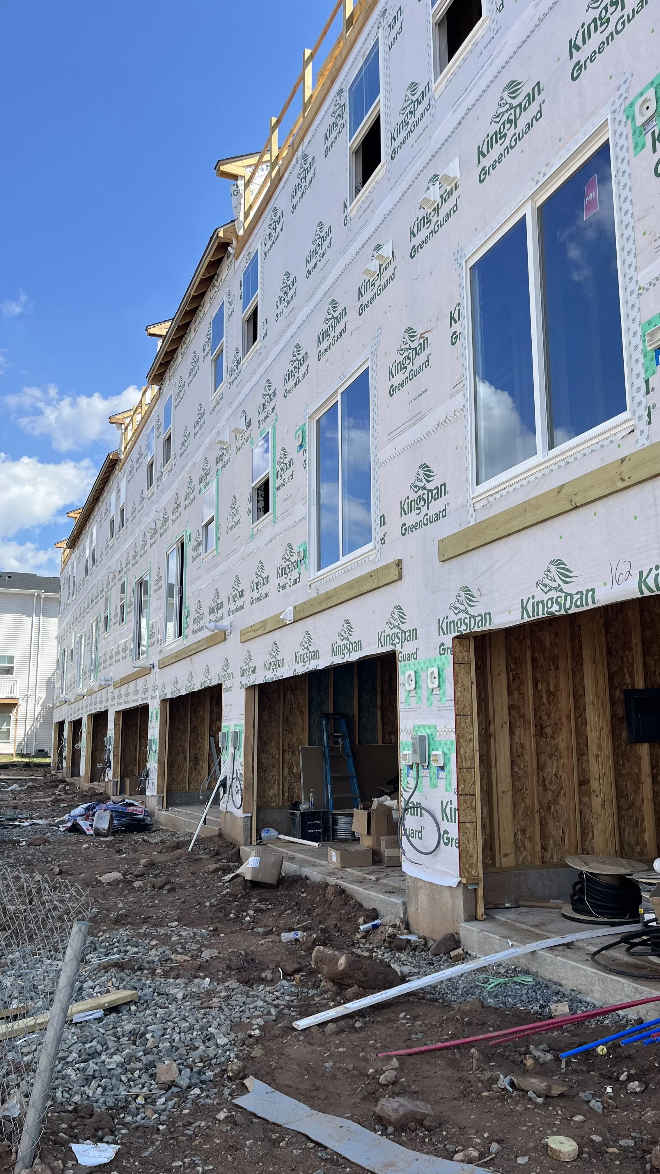 Construction site of a multi-story residential building with exterior insulation and windows installed. The building is unfinished with exposed framing and ground work, with blue sky and some clouds in the background.
