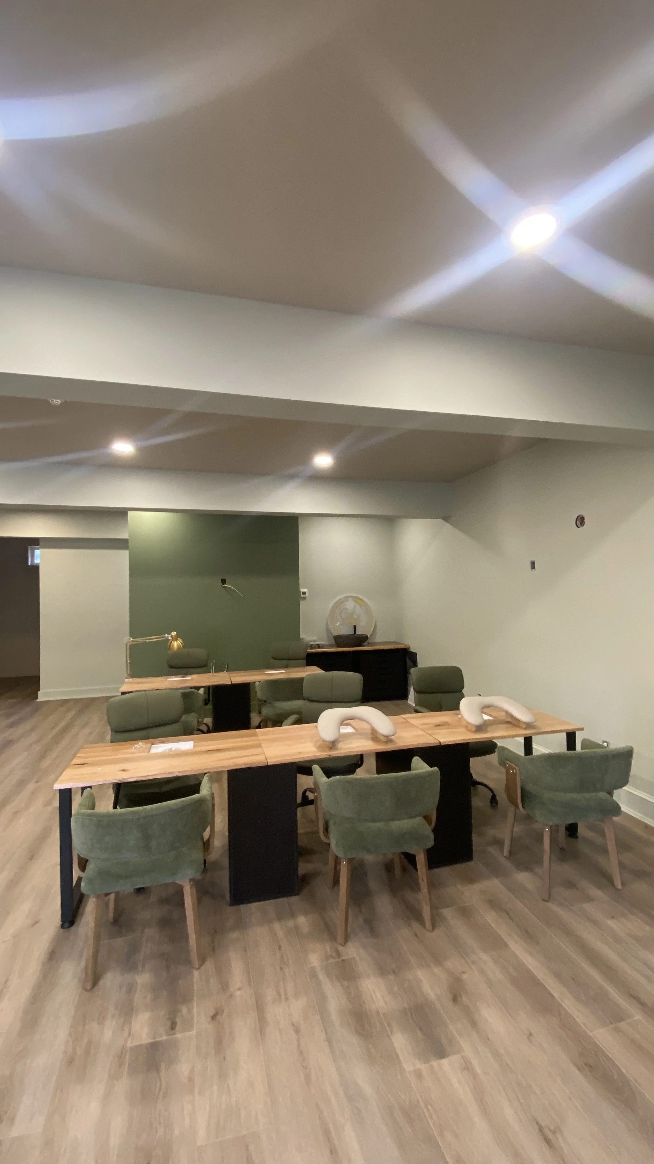 A room with wooden tables and green chairs arranged around them. The room has wood flooring, green and beige walls, and is illuminated by ceiling lights.