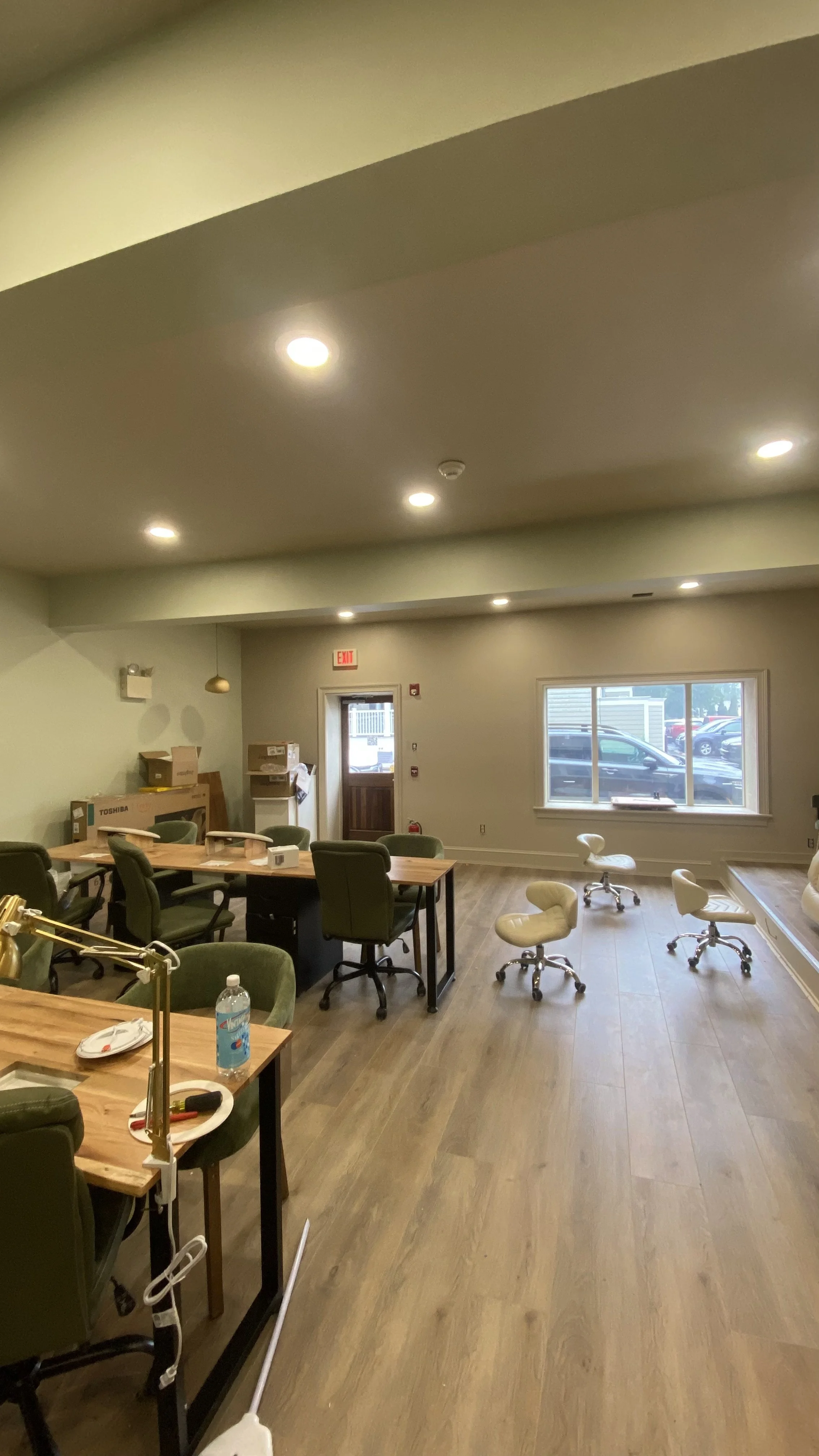 Office or meeting room with a wooden table, chairs, and stools, window showing parked cars outside, and some cardboard boxes near the door.