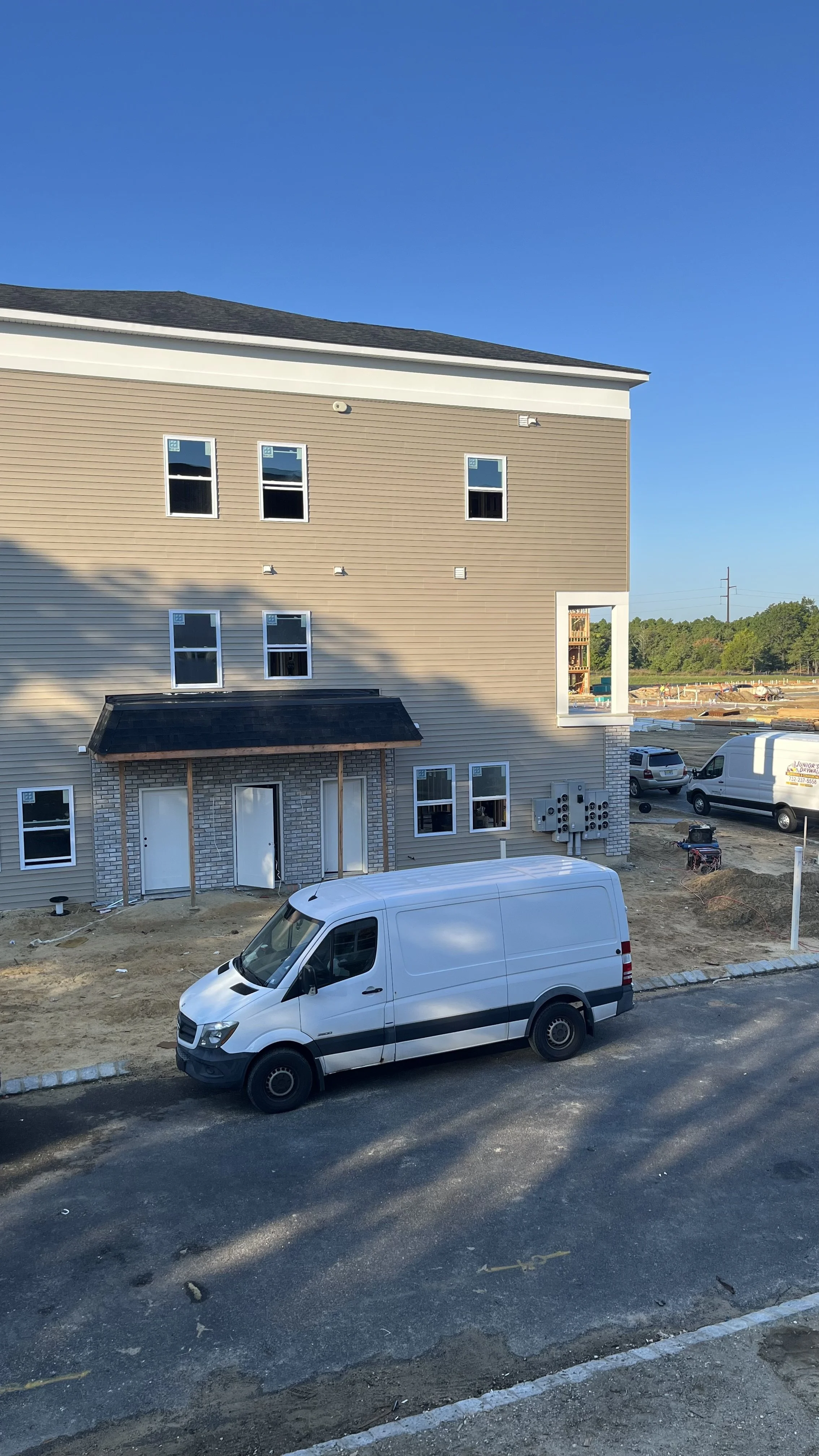 A multi-story residential building under construction with a white van parked in front. The building has beige and brick exterior walls, with several windows installed. There are construction materials and equipment visible around the site, and a cle
