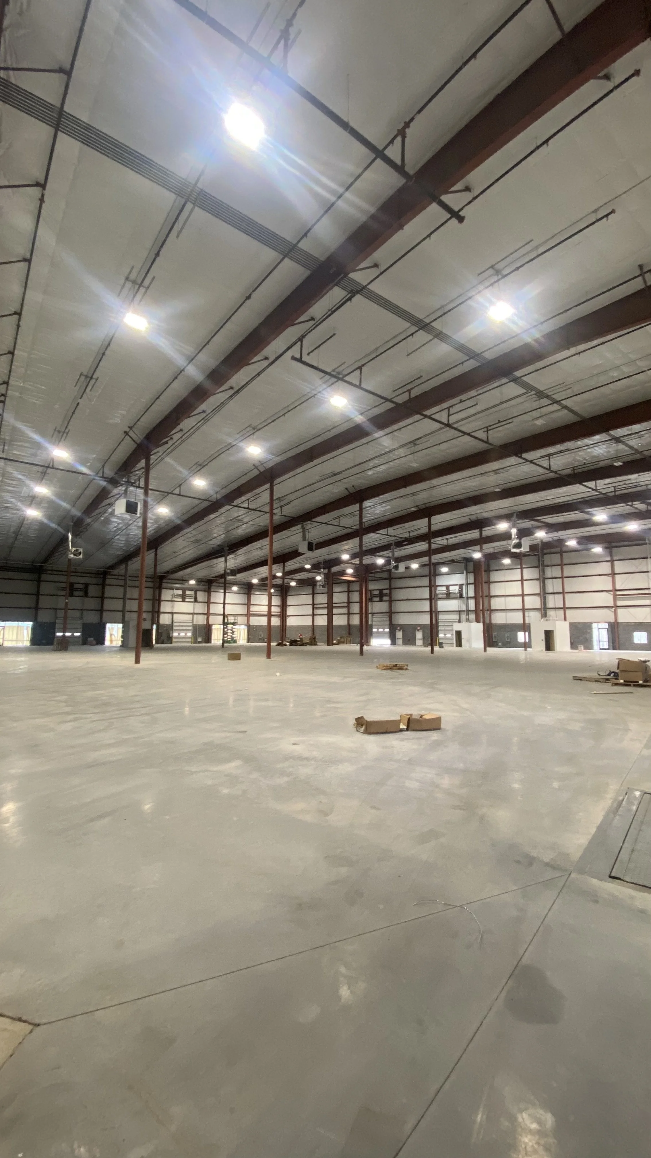 A large, empty warehouse with a concrete floor, high ceilings, steel beams, and bright overhead lighting. There are a few scattered boxes and pallets on the floor.