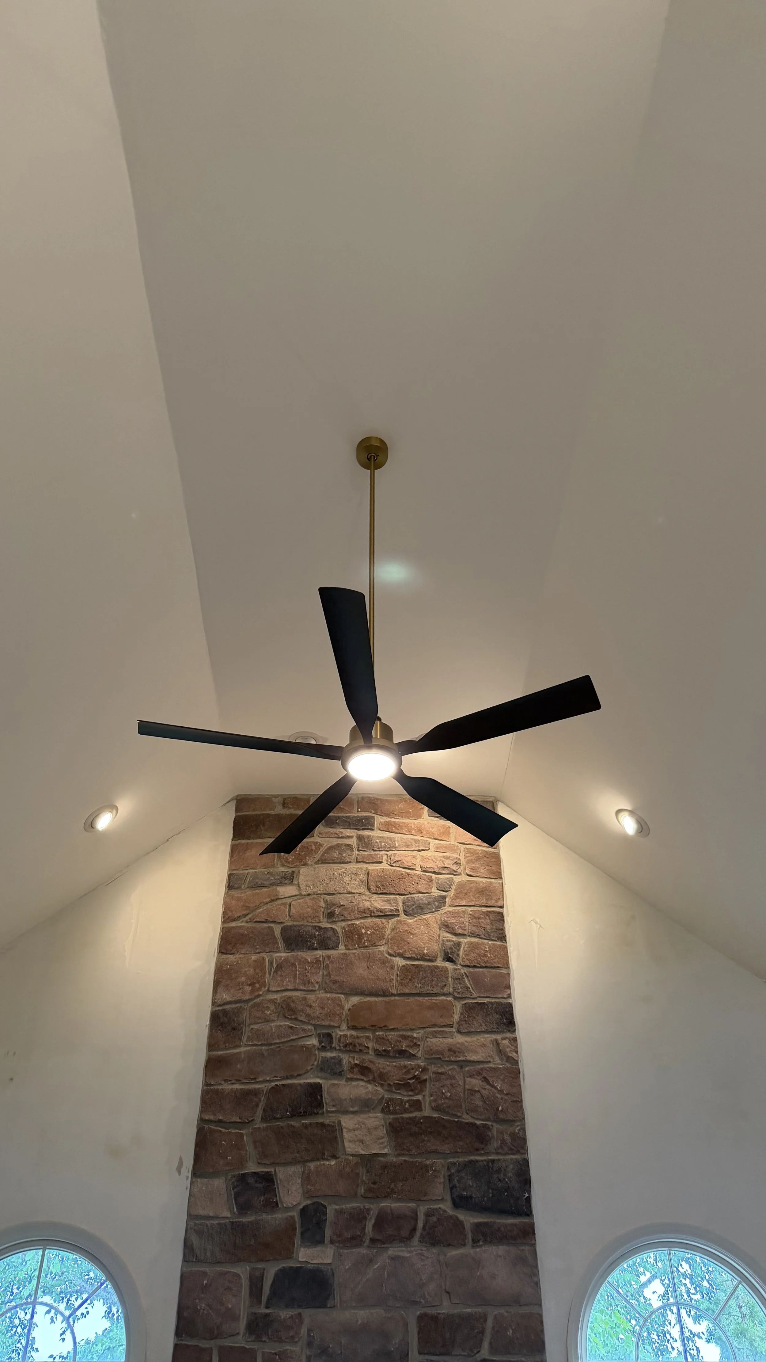 Ceiling fan hanging from a gold mount on a white ceiling, with a brick fireplace below and two arched windows with blue sky and greenery outside.