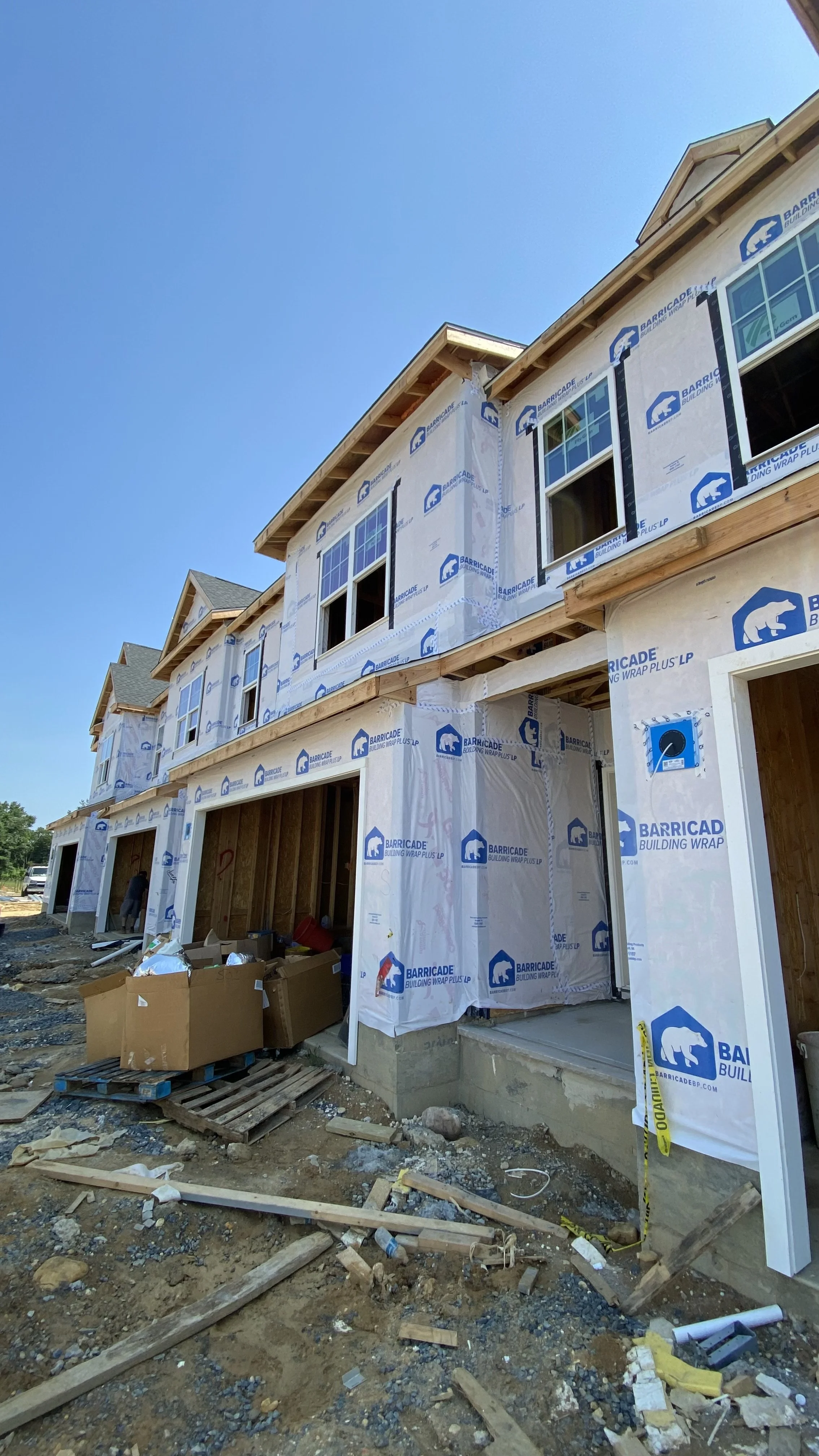 Construction site of a multi-family residential building with scaffolding and building wrap on the exterior walls, showing an early stage of construction with open garage spaces and construction materials on the ground.