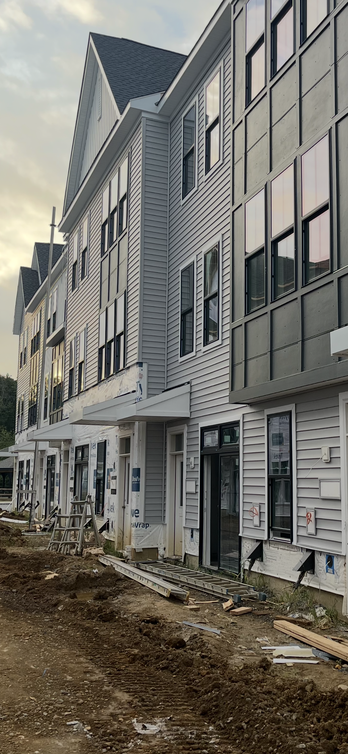 Construction site of a multi-story residential building with siding and large windows, construction materials and dirt ground in the foreground.