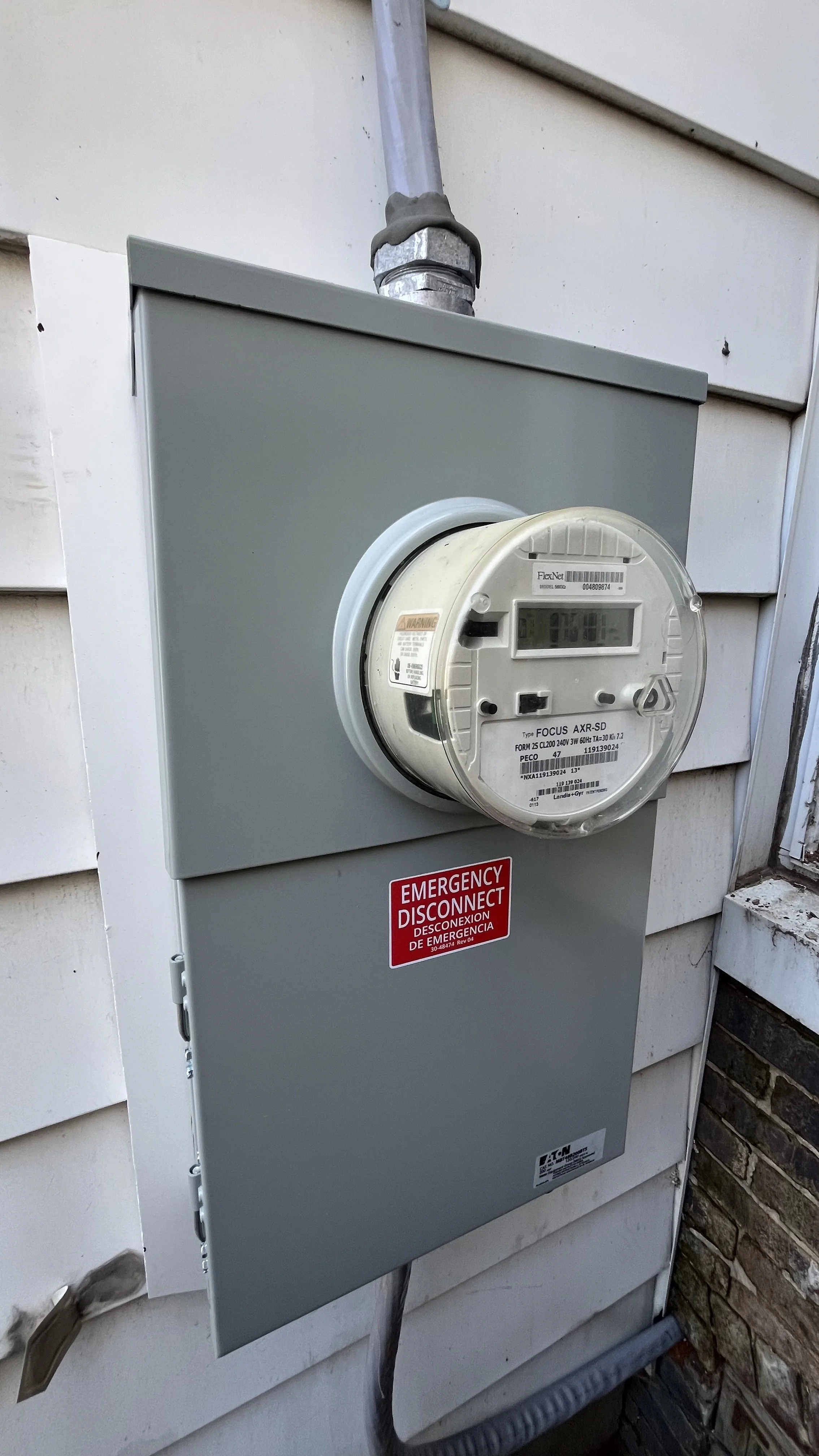 Electric meter mounted on an outdoor gray box with a red sticker indicating emergency disconnect, attached to a house with white siding and some brickwork.