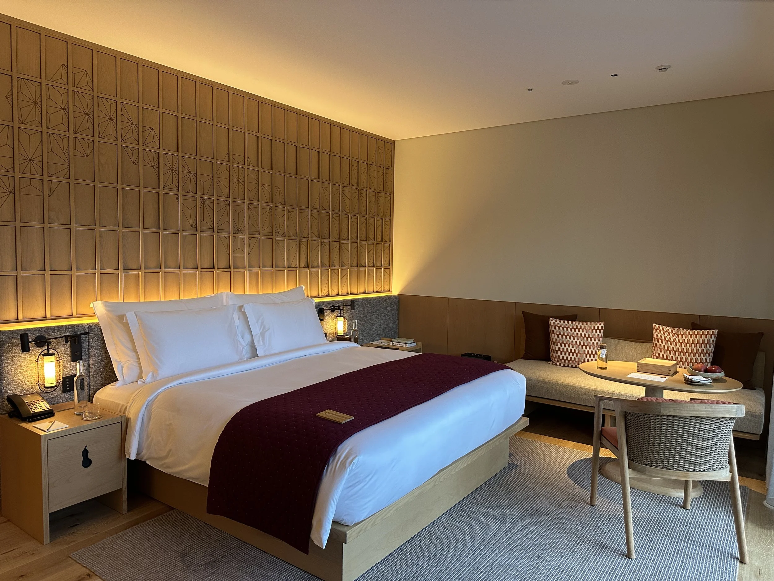 A bedroom with a large bed, white linens, a purple bed runner, and pillows. There is a wooden headboard with geometric patterns, nightstands with lamps, and a sitting area with a table, chairs, and decorative pillows.