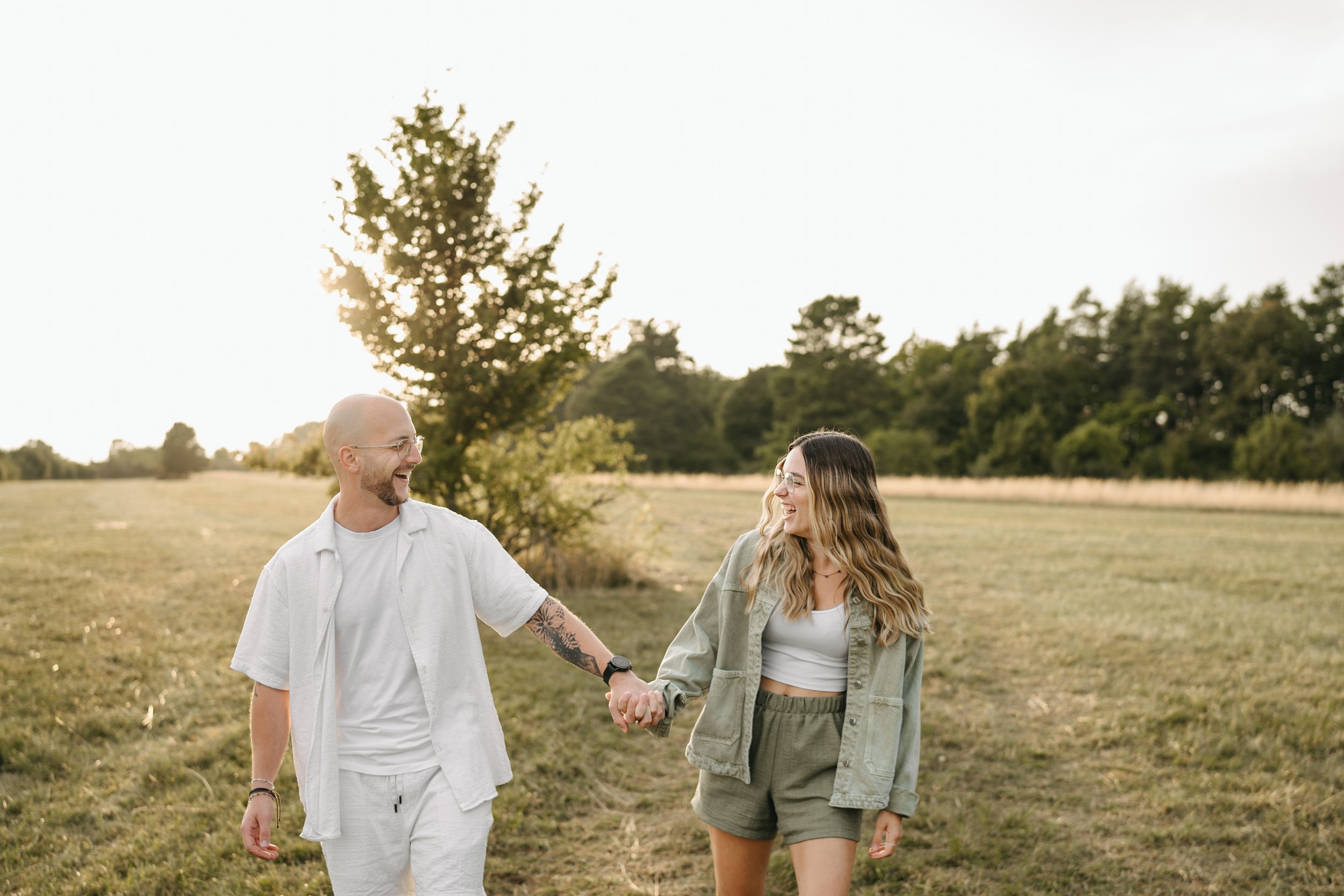 Ein Mann und eine Frau spazieren lächelnd in einem offenen Feld bei Sonnenuntergang, während sie sich an den Händen halten.