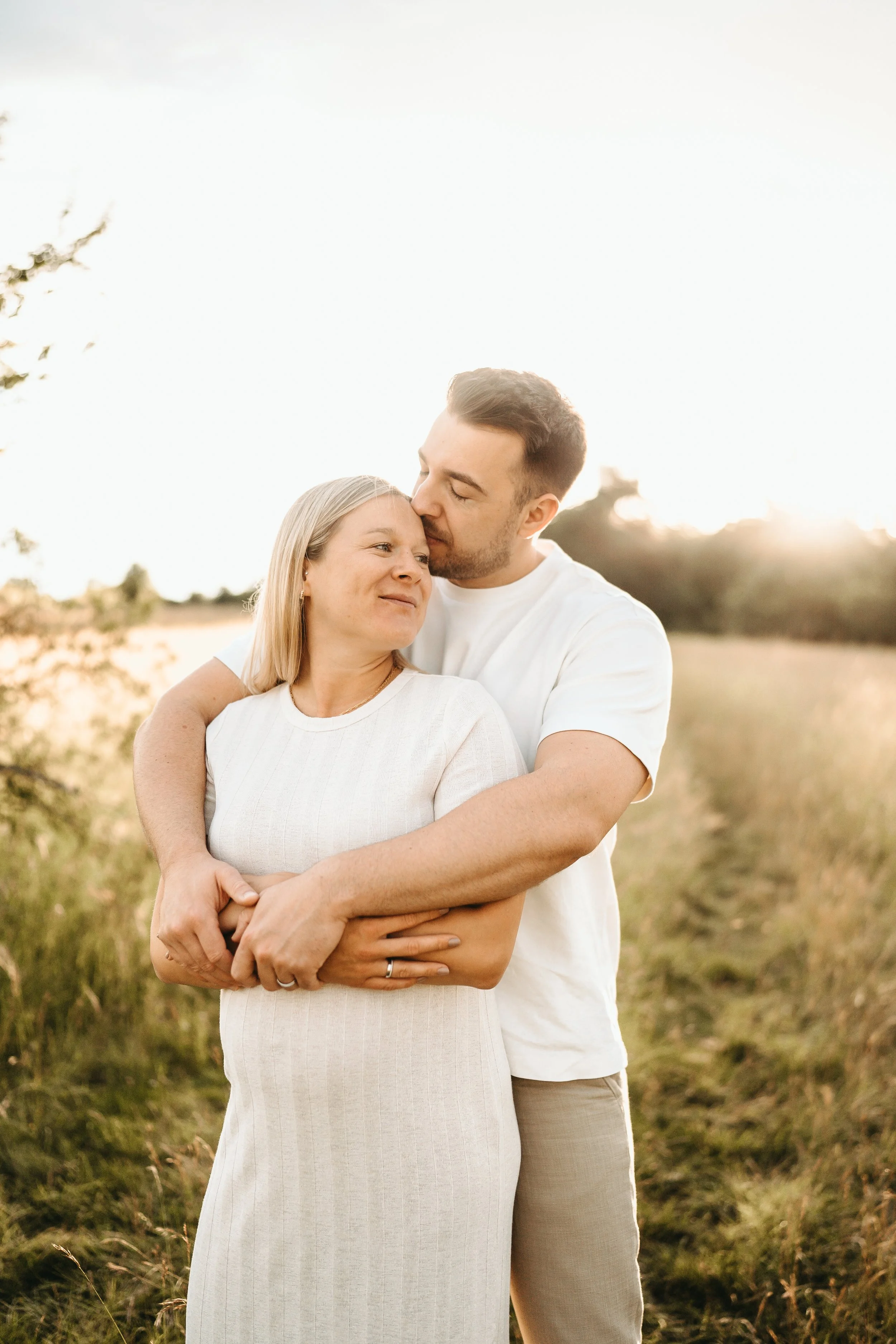 Ein Paar steht in einer Wiese bei Sonnenuntergang, umarmt sich liebevoll und schaut sich an.