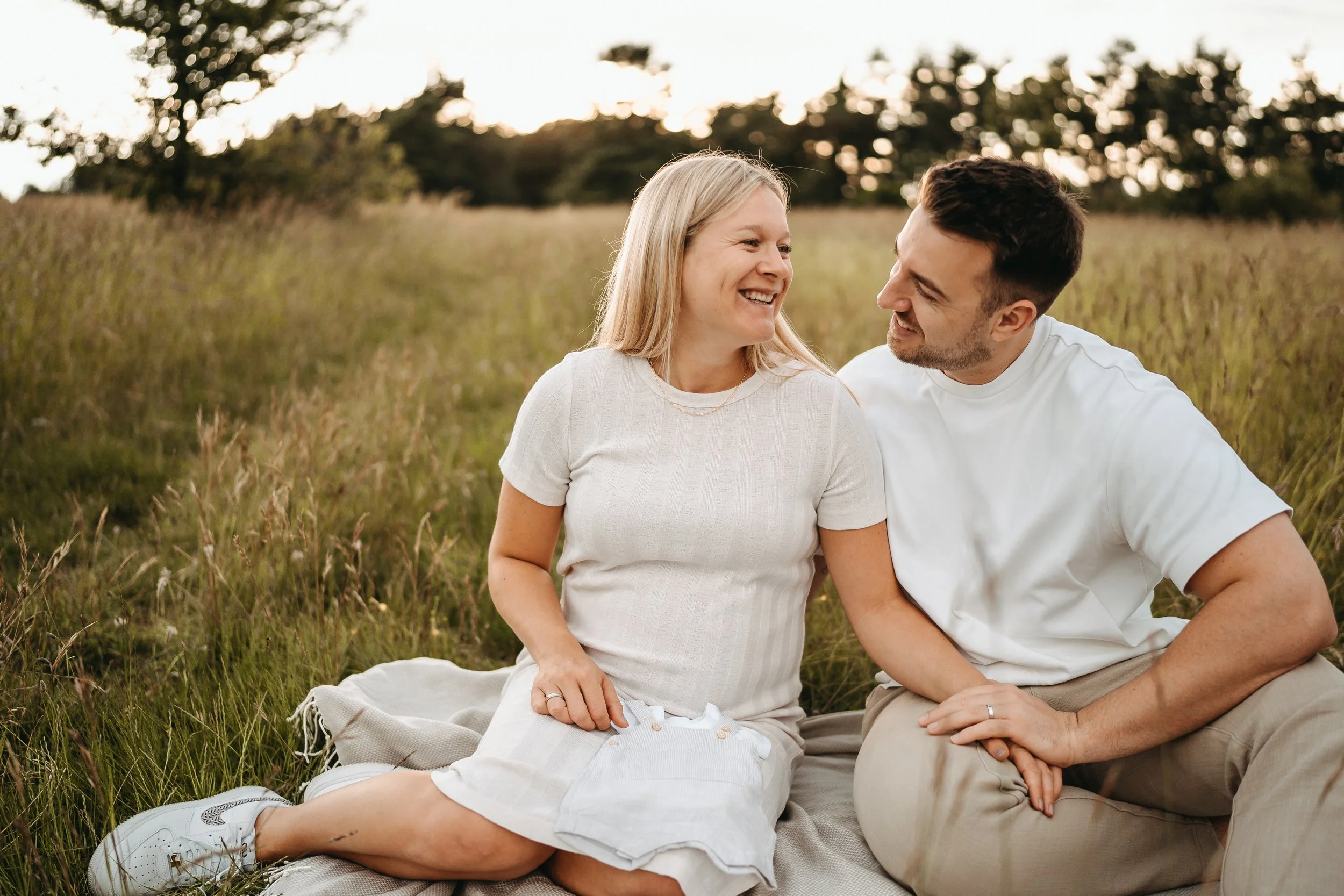 Ein Mann und eine Frau sitzen auf einer Decke in einem Feld bei Sonnenuntergang und lachen miteinander.