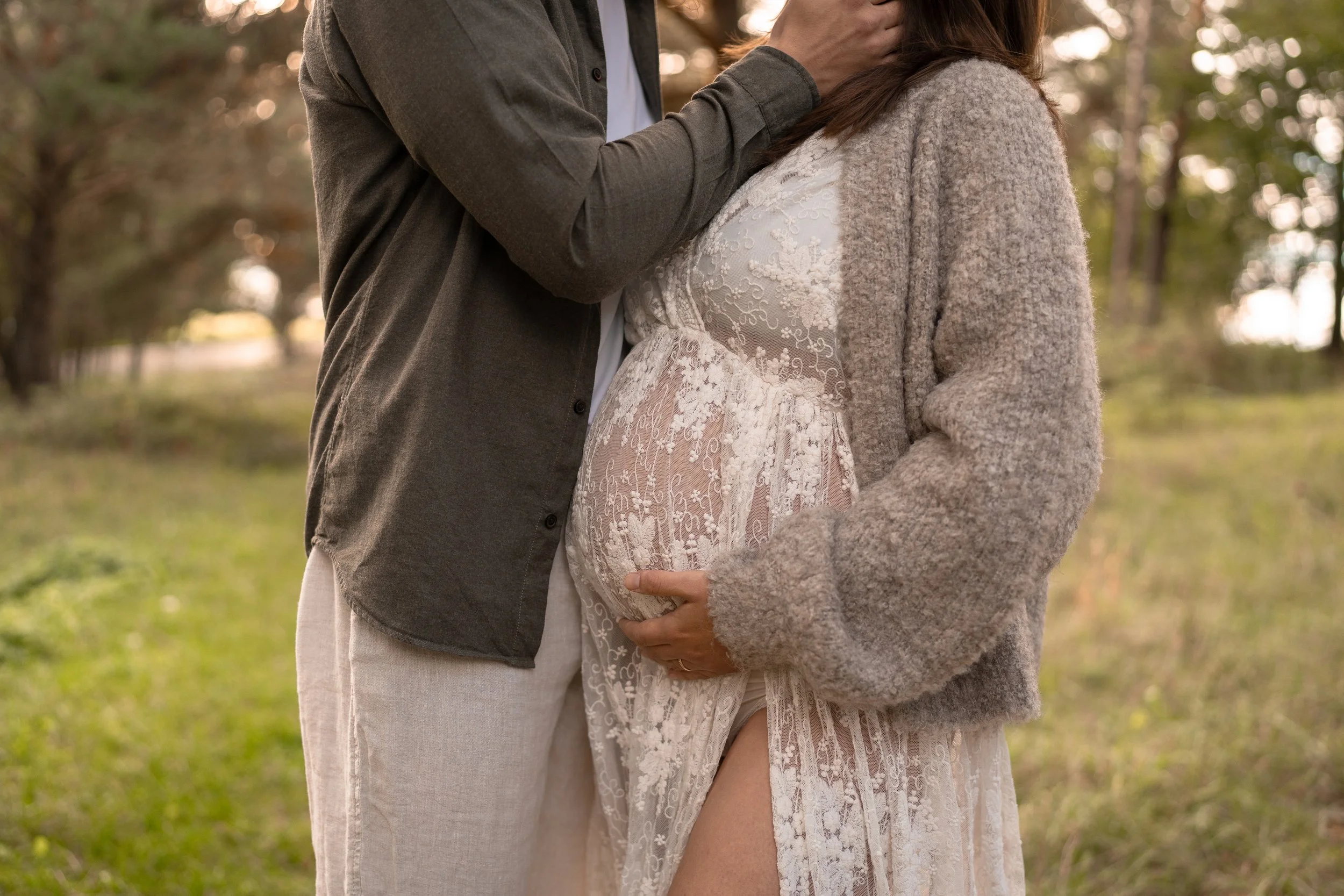 Ein schwangere Frau in einem eleganten, durchsichtigen Spitzenkleid, die in einem Park steht, umgeben von Bäumen, während ihr Partner sie liebevoll berührt.